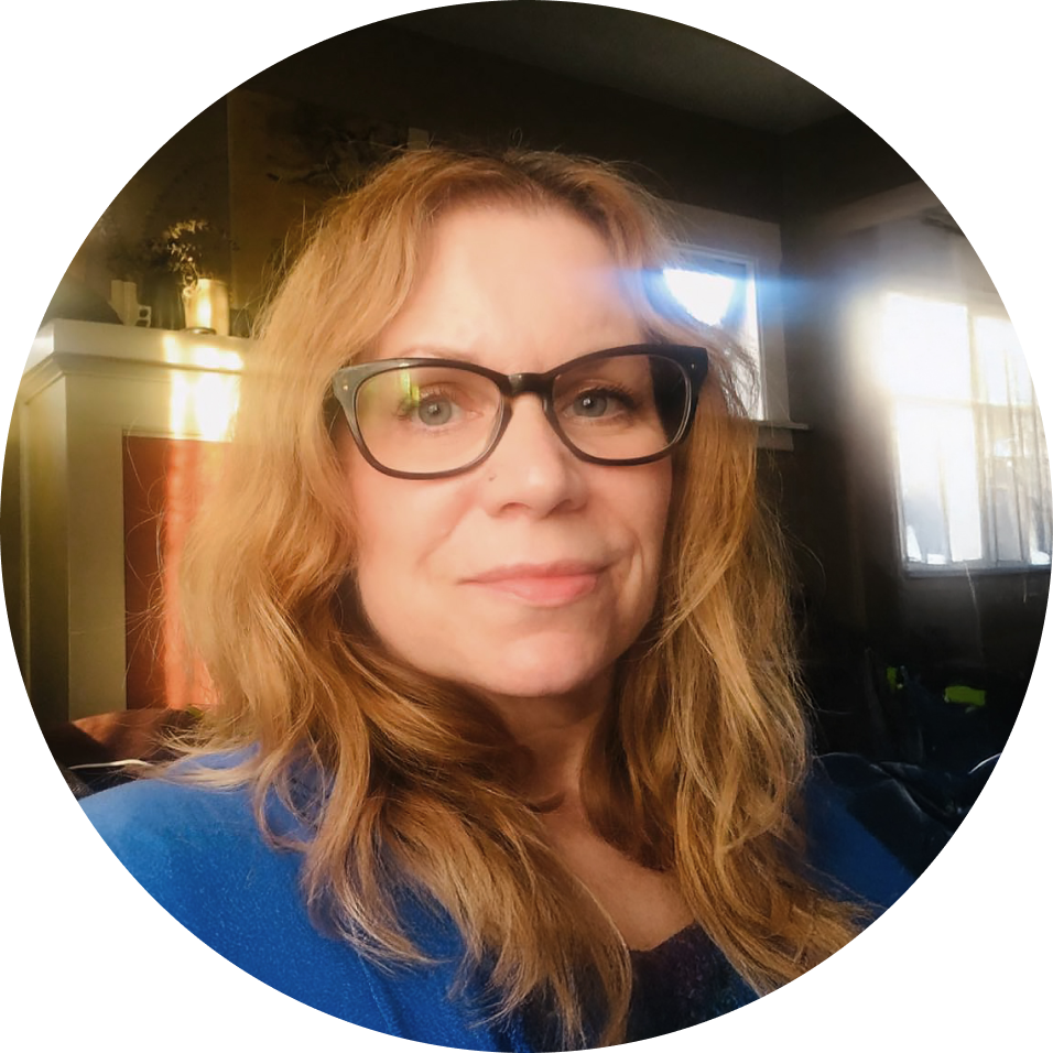 A woman with red hair and glasses smiling indoors, with sunlight coming through a window behind her.