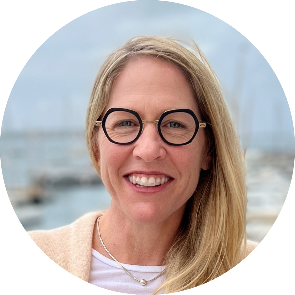 Close-up of a woman with blonde hair, glasses, smiling, with water and a boat in the background.