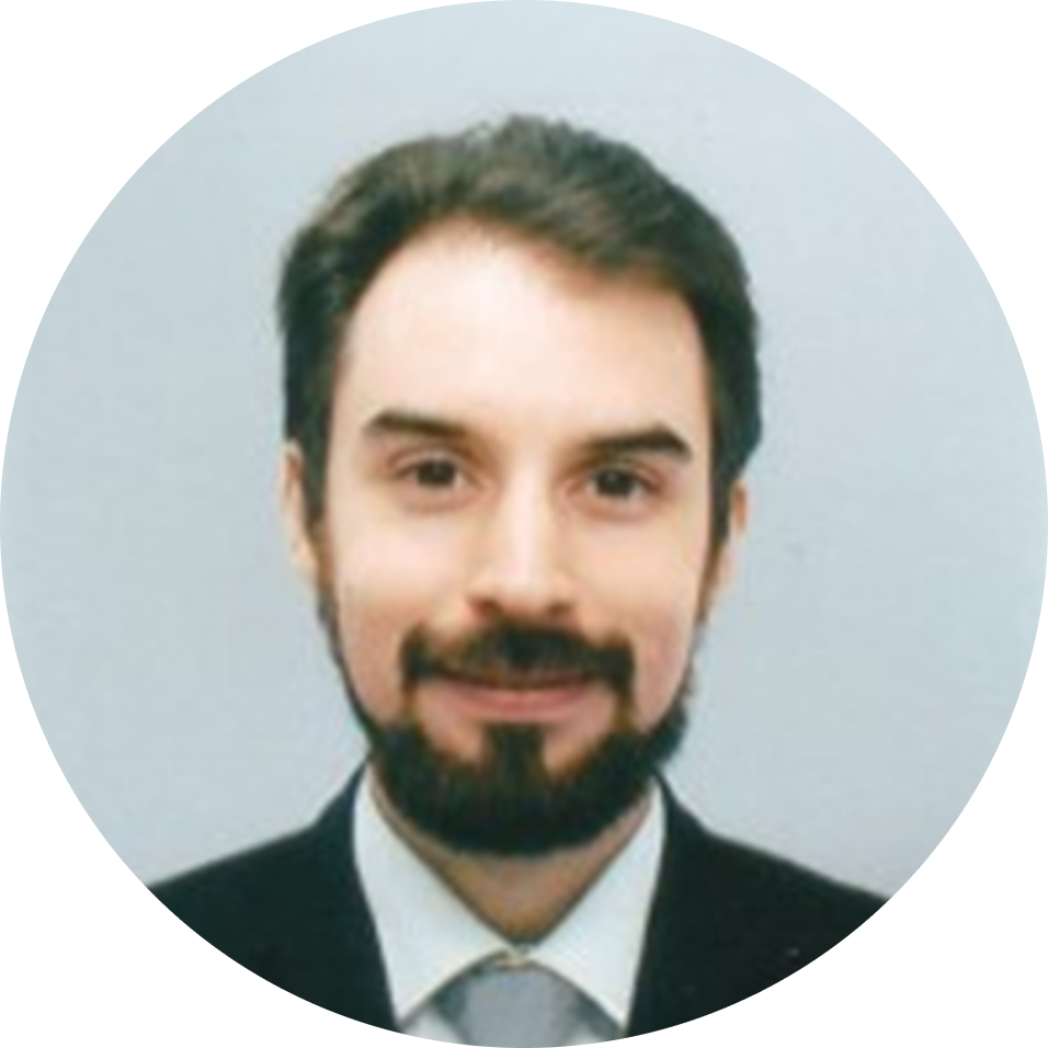 Headshot of a man with dark hair and a beard, wearing a suit and tie, smiling against a light background.