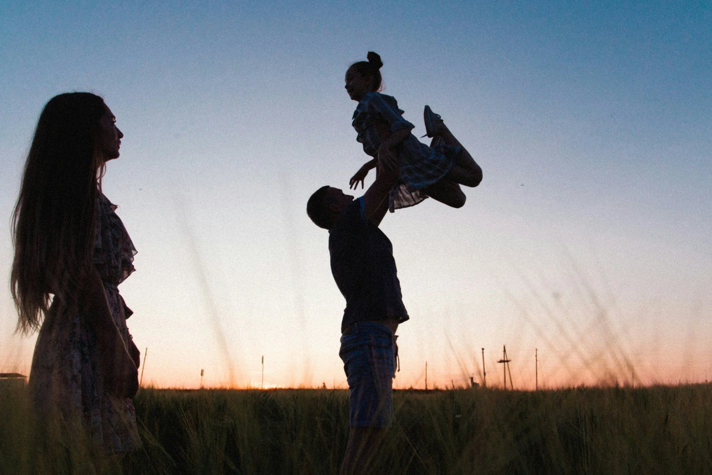 Familie beim Spielen im Feld bei Sonnenuntergang, Mann hebt ein Kind hoch, während eine Frau zuschaut.