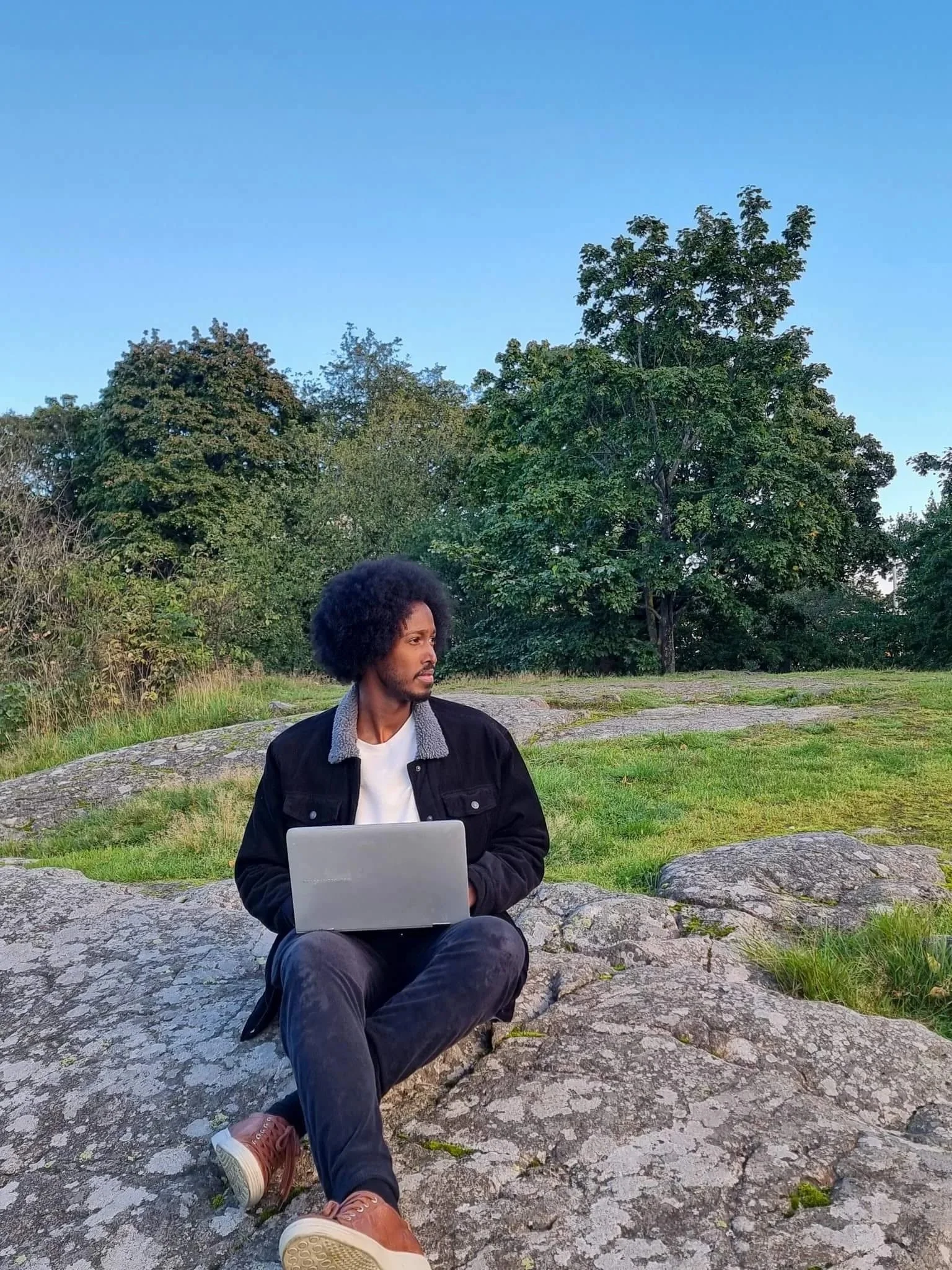Image of the squarepace web design studio owner Daahir Abdulle sitting on a hill while hlding his laptop on his lap as he looks to his left (viewers right).