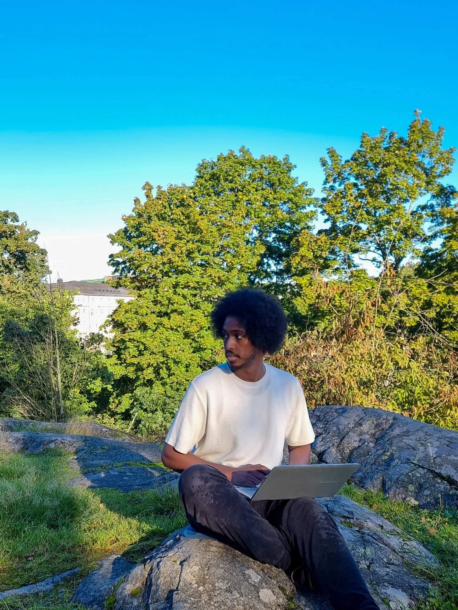 Image of Daahir Abdulle sitting ona hill with his laptop on his lap, wearing a white shirt. Looking to his right (viewer's left)
