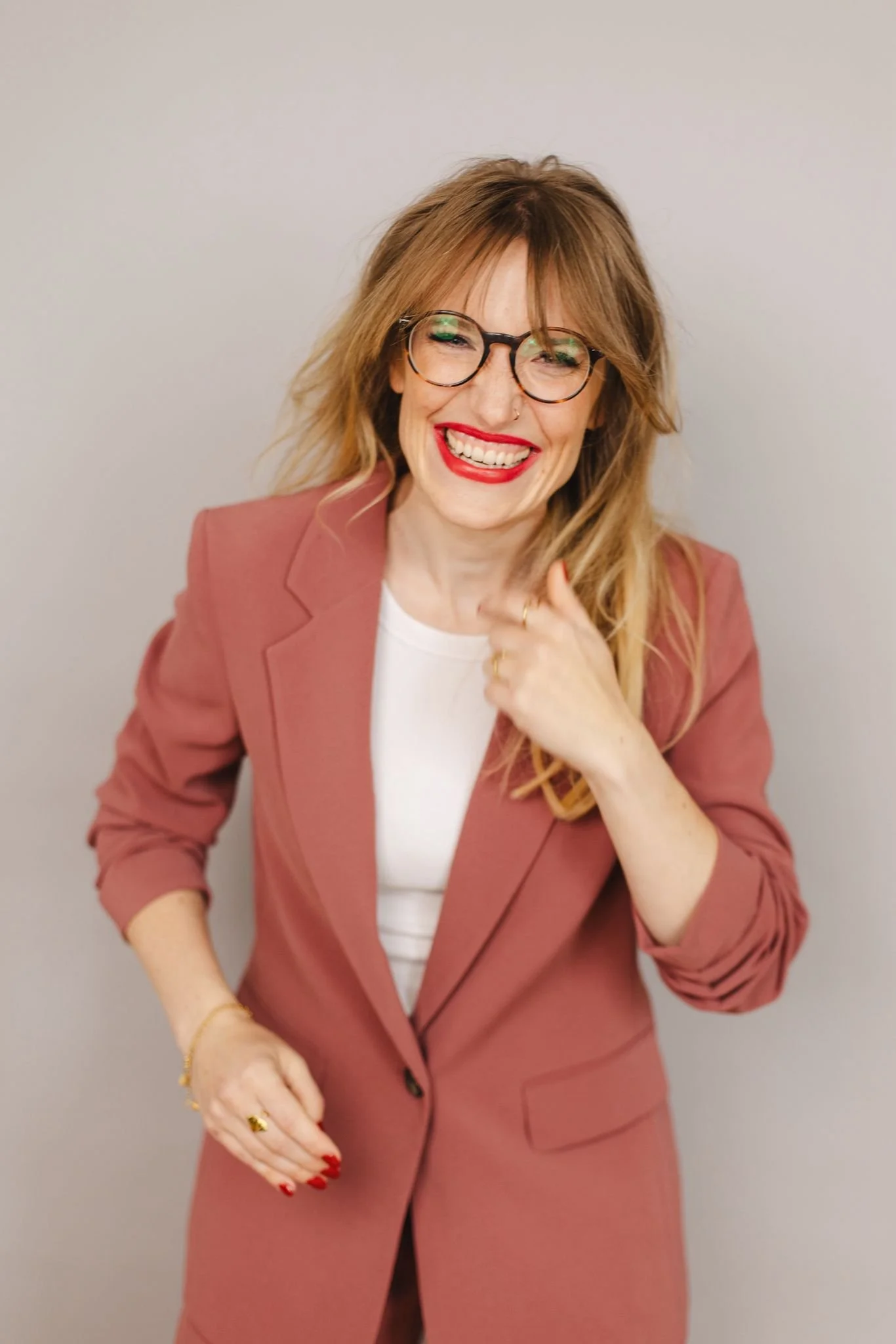 Lächelnde Frau mit Gläsern, rotem Lippenstift in rosa Blazer vor neutralem Hintergrund.