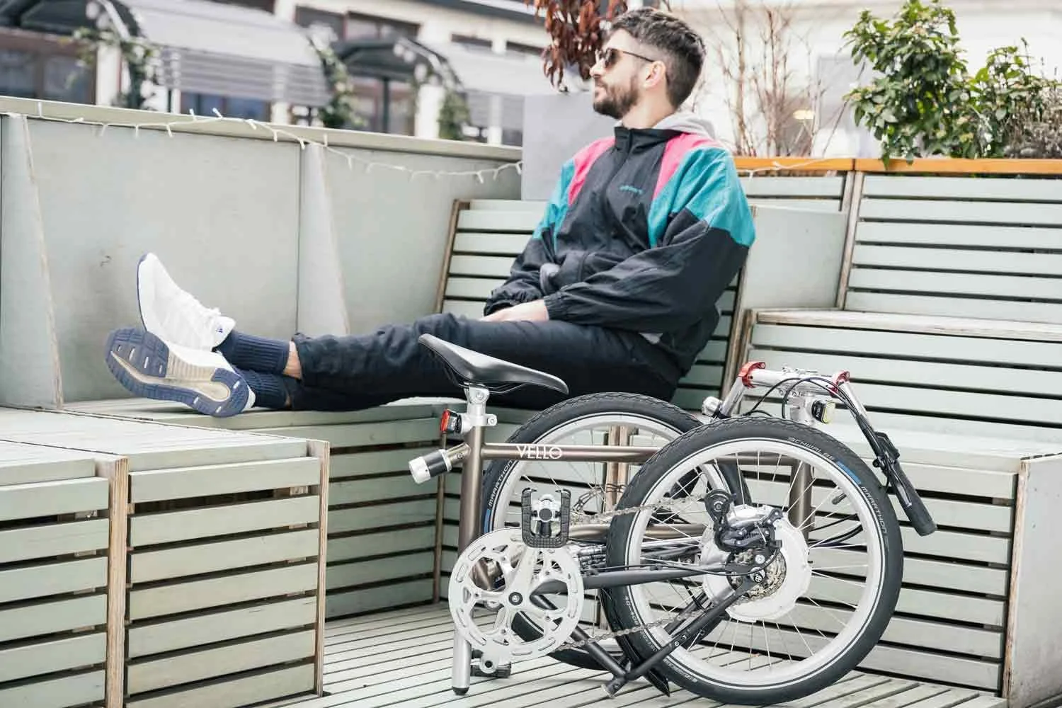 Un homme avec des lunettes de soleil et un casque de vélo, assis sur un banc en bois, avec un vélo électrique pliable à côté, dans un espace extérieur urbain.