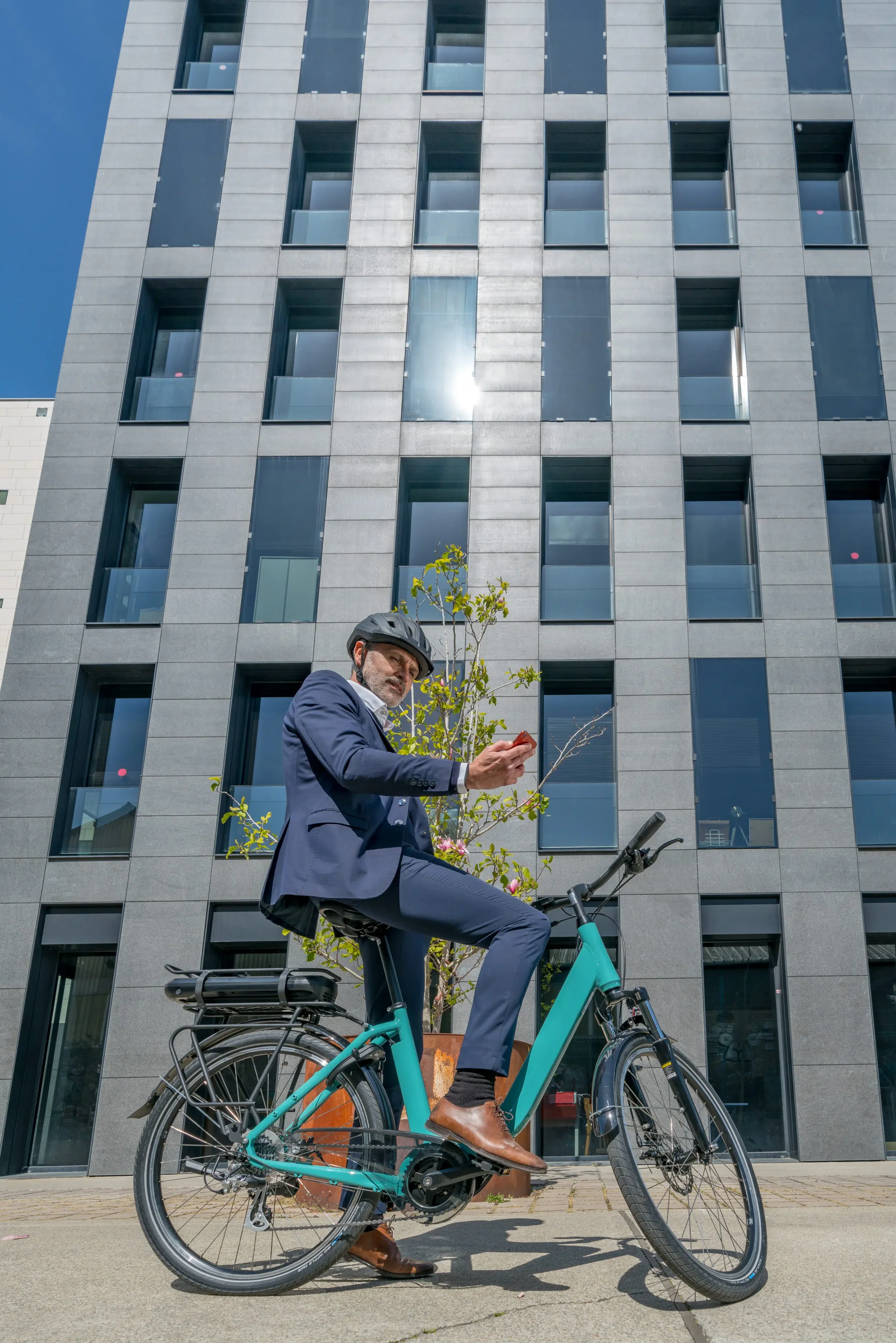 Un homme en costume et casque de vélo, assis sur un vélo électrique turquoise, regardant son téléphone, devant un bâtiment moderne avec plusieurs fenêtres, arbres et ciel bleu.