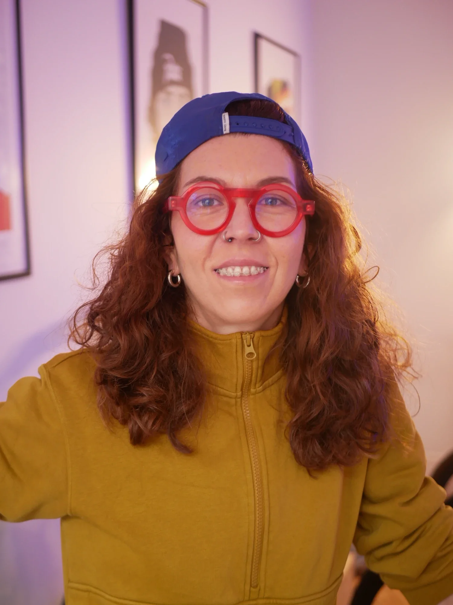 Jeune femme avec lunettes rouges, casquette bleue, veste jaune, souriant dans un intérieur avec des œuvres d'art sur le mur.