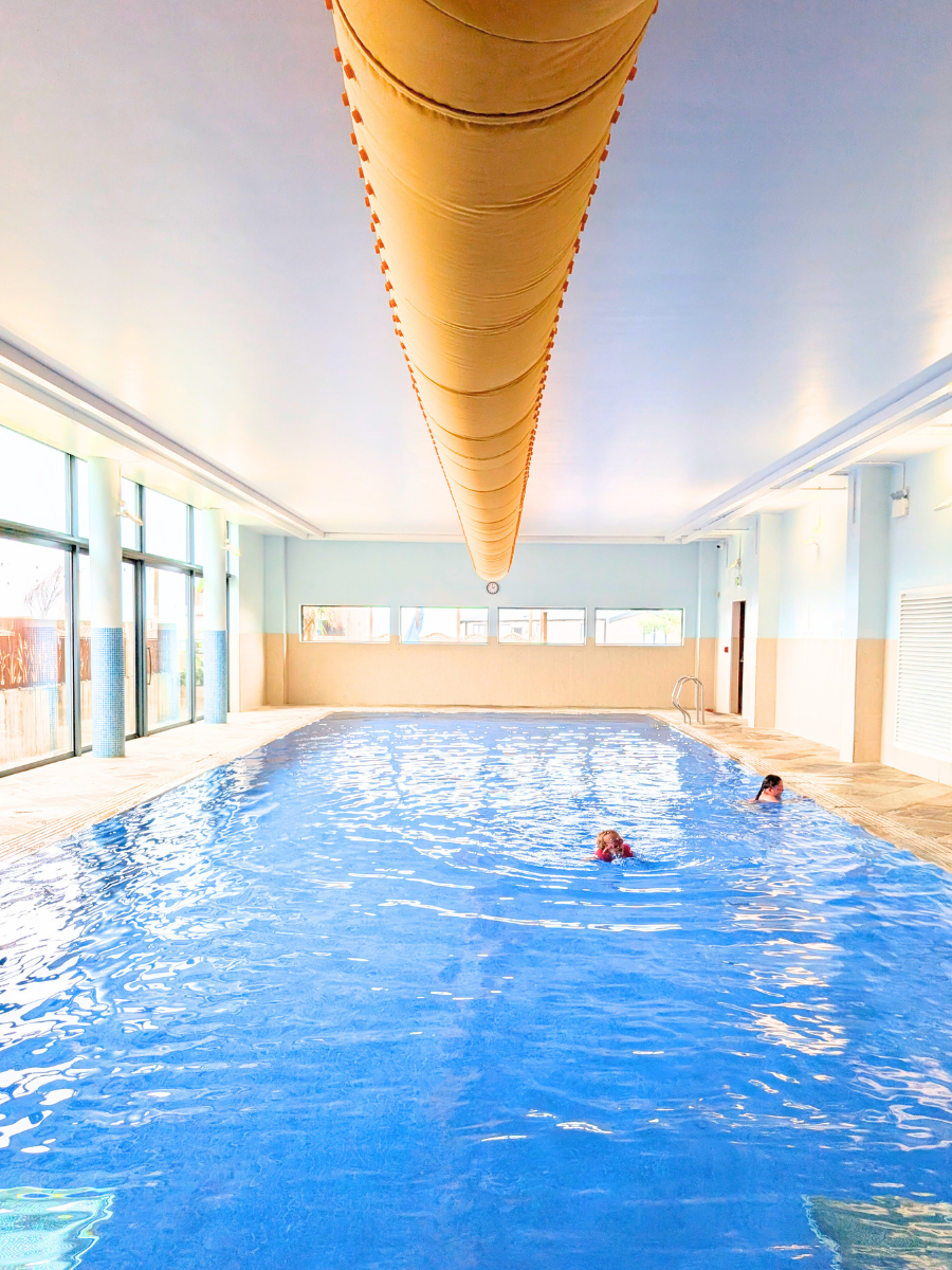 Indoor swimming pool with two people swimming, large windows on the left, a yellow pool cover hanging from the ceiling, and a door on the far right.