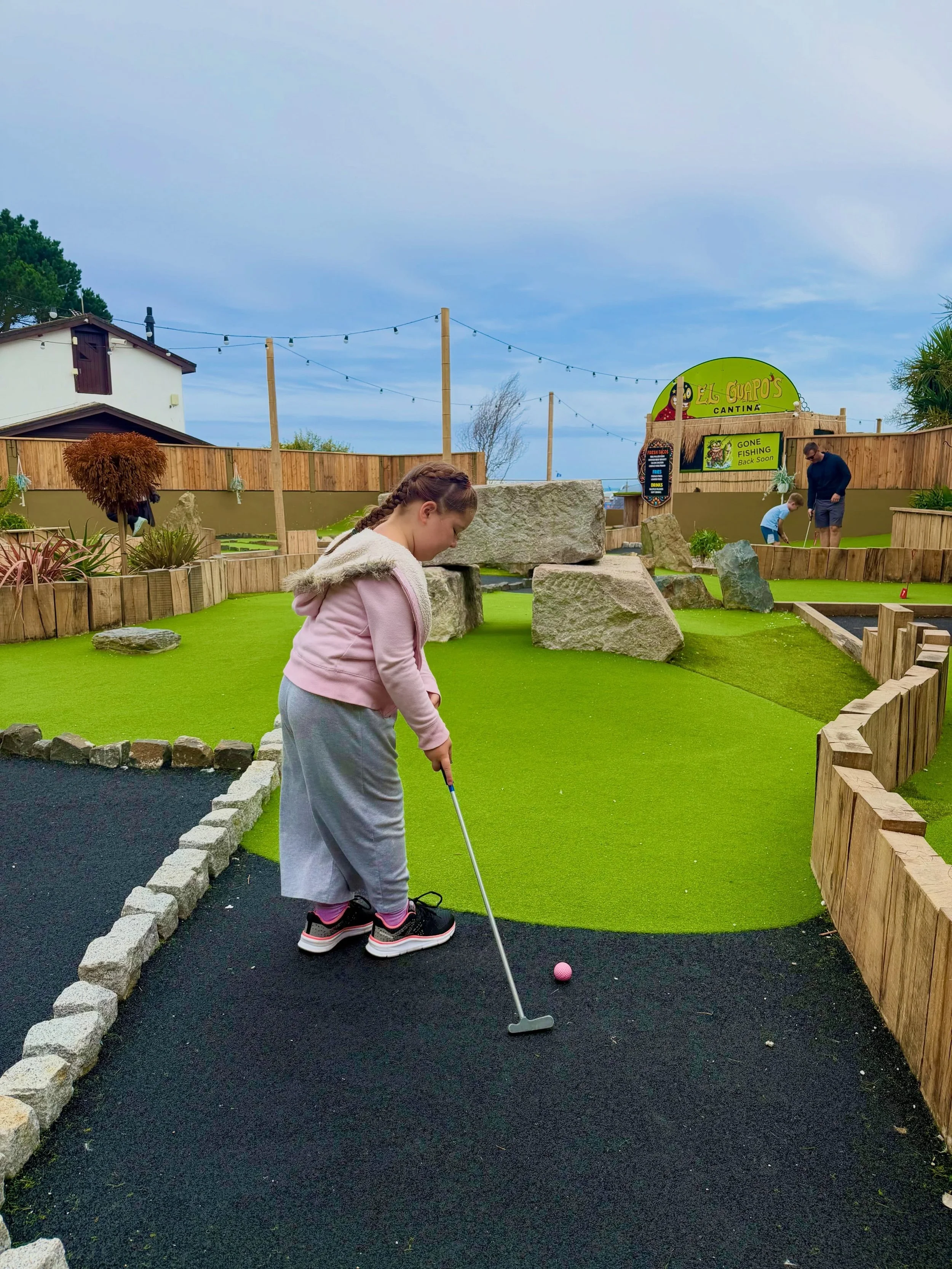 A young girl with braided hair and gray sweatpants playing mini-golf on an artificial green turf course with large rocks and wooden border. In the background, a man and a boy are playing, and there is a wooden fence, a sign for a cantina called El Guapo's, and string lights hanging above.