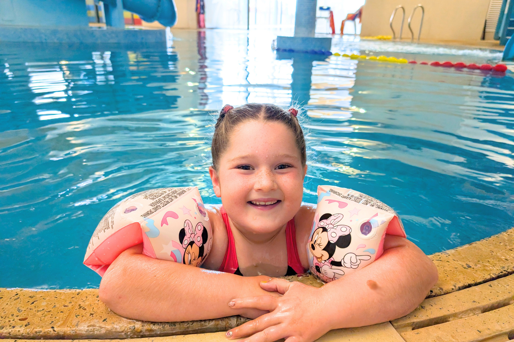 A young girl with red hair in pigtails, wearing Minnie Mouse-themed arm floats, smiling in a swimming pool with her arms resting on the pool edge and water around her.