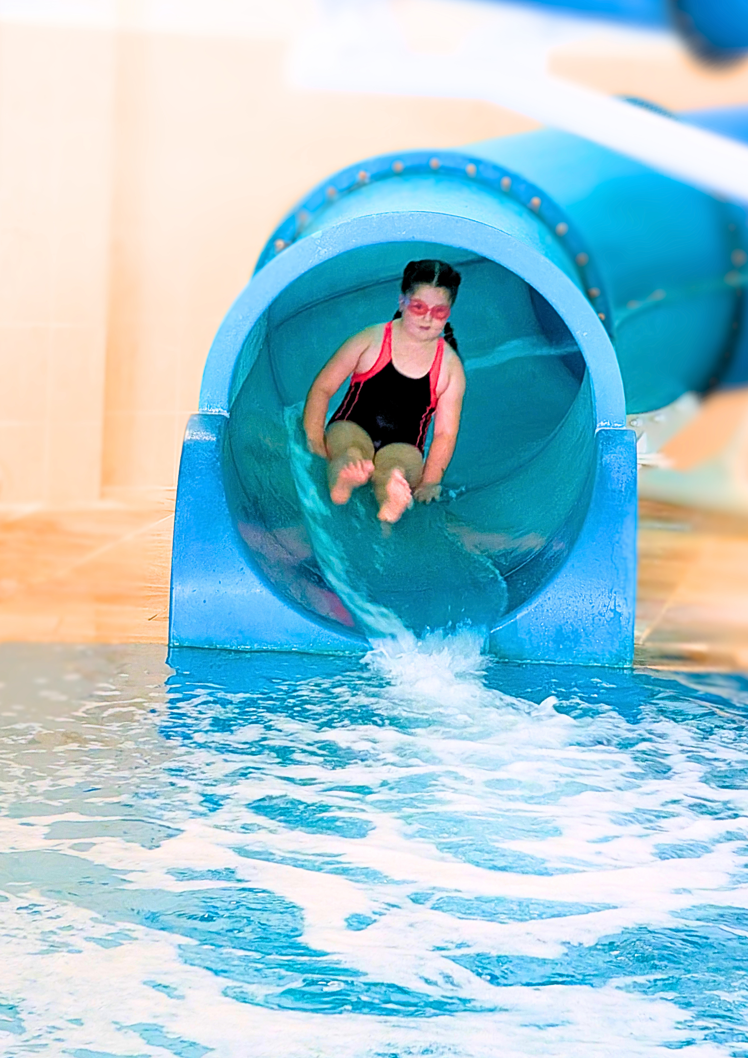 Child girl in a black swimsuit with pink straps and pink swim goggles exiting a blue water slide into a swimming pool.