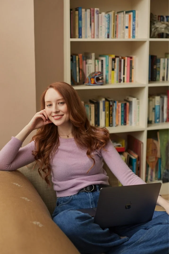 A young woman with long red hair, wearing a pink sweater and blue jeans, relaxing on a beige sofa with a laptop on her lap in a room with a bookshelf filled with books.