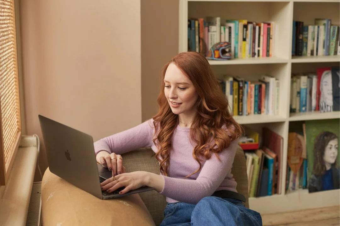A young woman with long red hair working on a laptop, sitting on a couch near a window with a bookshelf in the background.