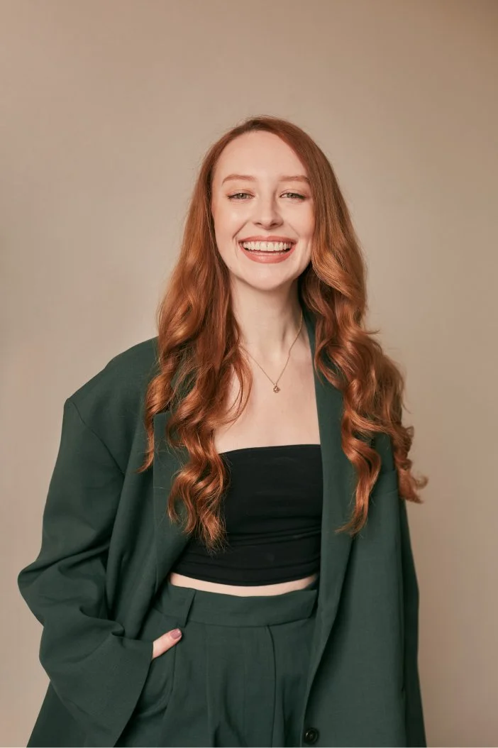 Woman with long curly red hair smiling, wearing a dark green suit and black crop top, standing against a beige background.
