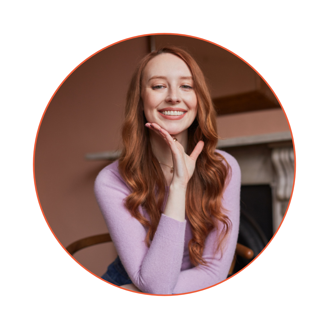 A smiling woman with long red hair, wearing a lavender sweater, sitting indoors with a fireplace in the background.