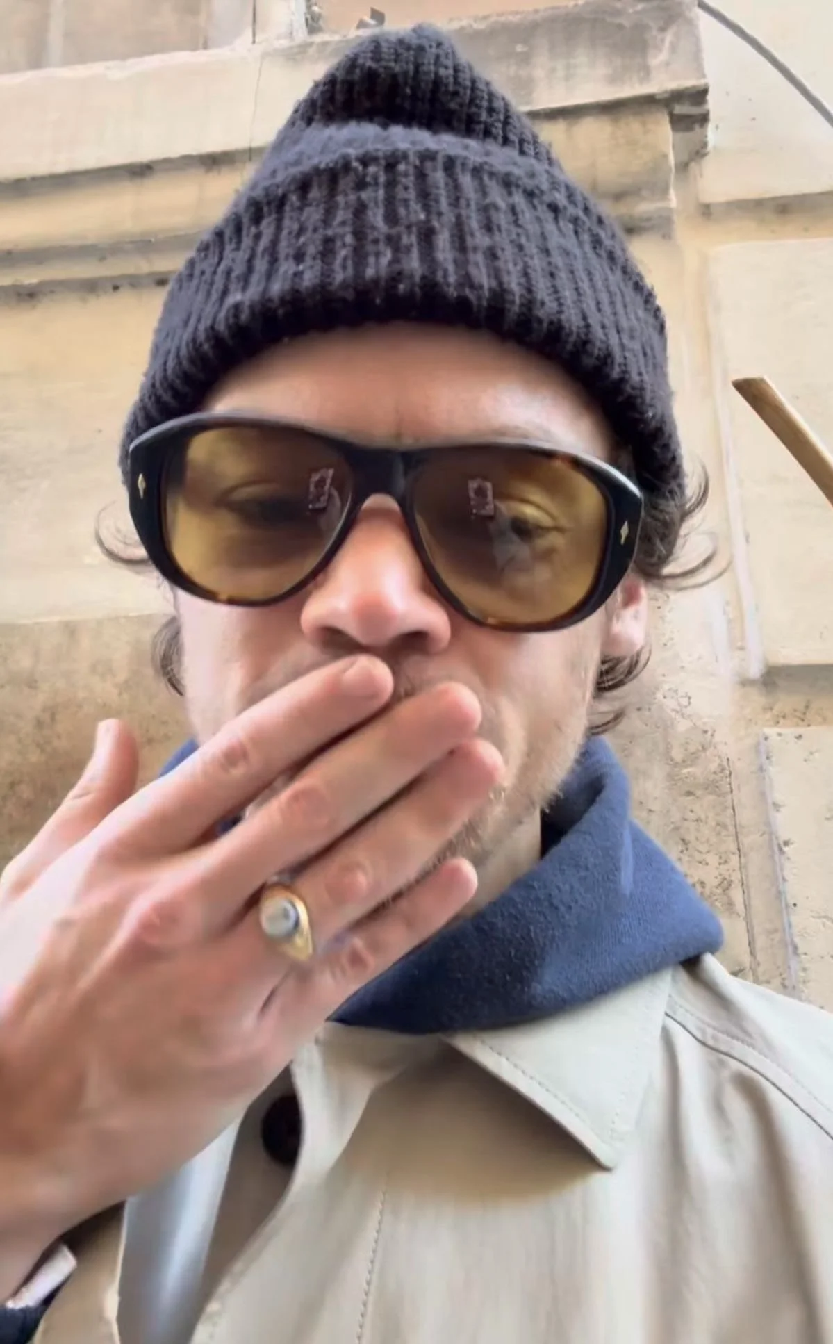 Harry Styles Junge trägt eine graue Jacke, einen schwarzen Strickhut, braune Sonnenbrille und eine Hand mit einem Ring hält vor den Mund.