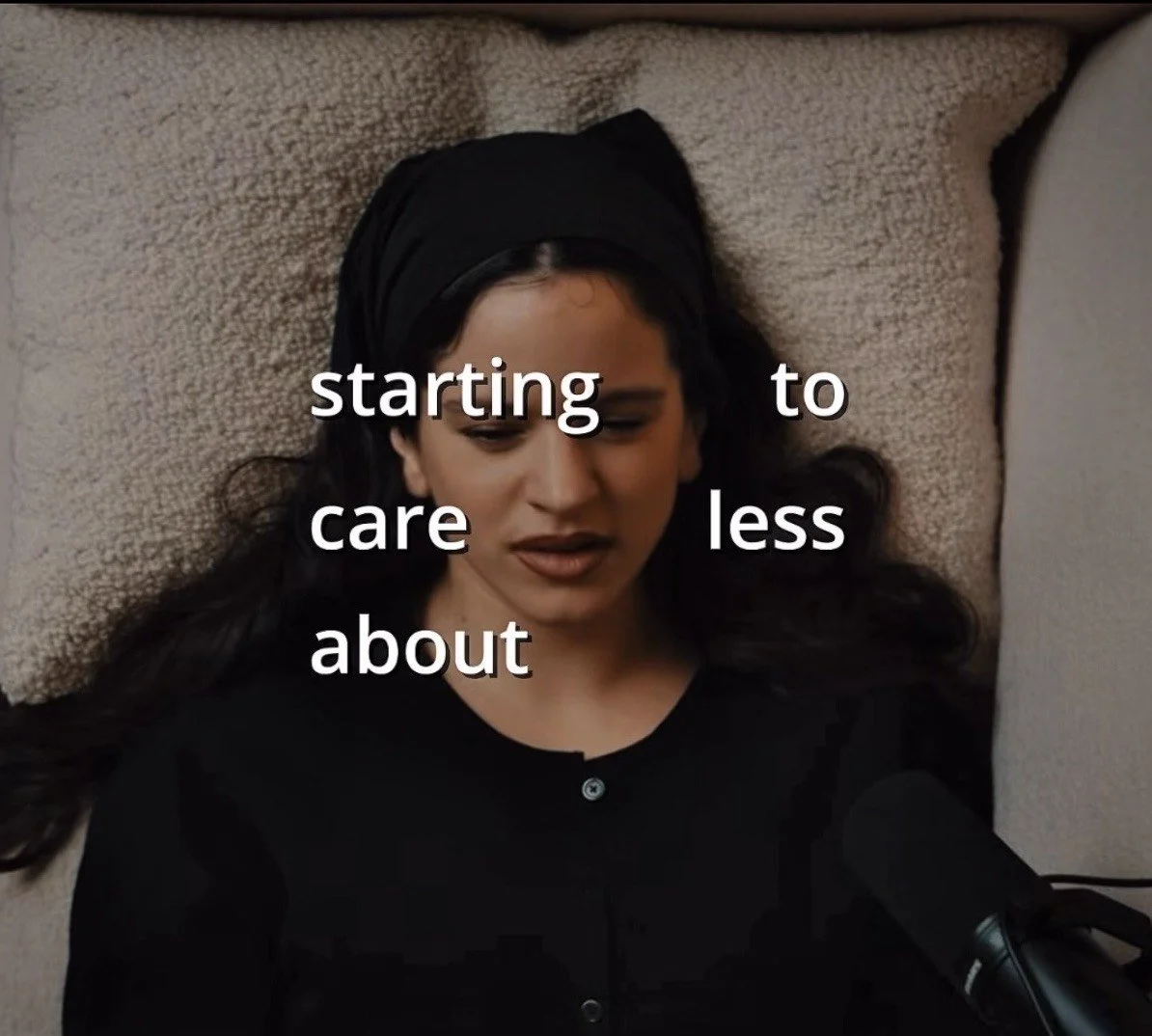 Rosalia La Perla Eine junge Frau mit dunklen Haaren, die auf einem Kissen liegt, trägt einen schwarzen Schal und schwarze Kleidung. Im Vordergrund befindet sich ein Mikrofon.