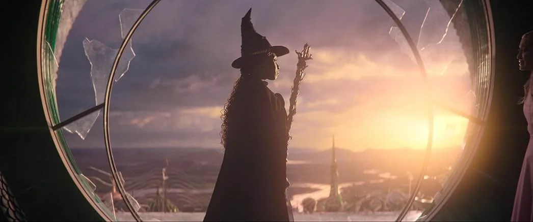 Wicked Universal Pictures Silhouette eines Zauberers mit Hut und Zauberstab vor einem Sonnenuntergangs-Hintergrund, gesehen durch ein zerbrochenes Fenster.