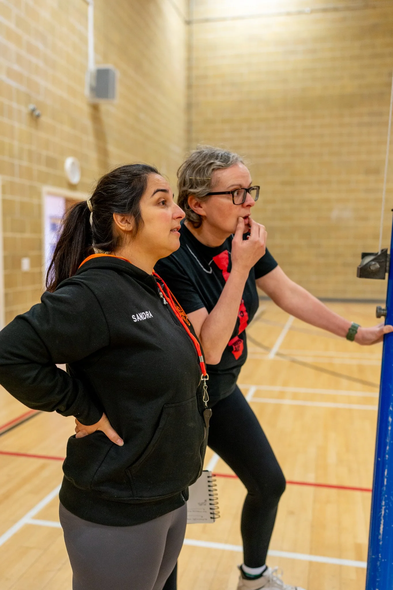 Two women standing side by side in a gymnasium, looking at a scoreboard or screen, with one woman wearing a black jacket with the name 'Sandra' on it and the other with glasses and short gray hair, both appearing focused.