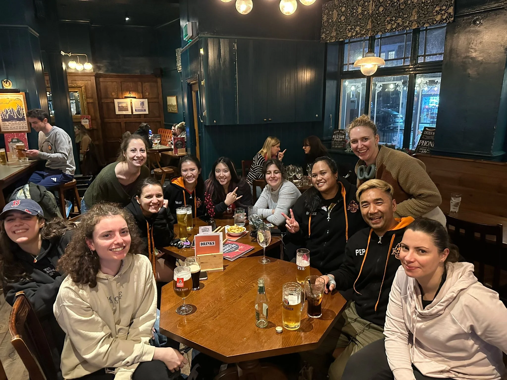 Group of people sitting around a table at a restaurant or bar, smiling and posing for the camera with drinks and food on the table, in a cozy, dimly-lit interior.