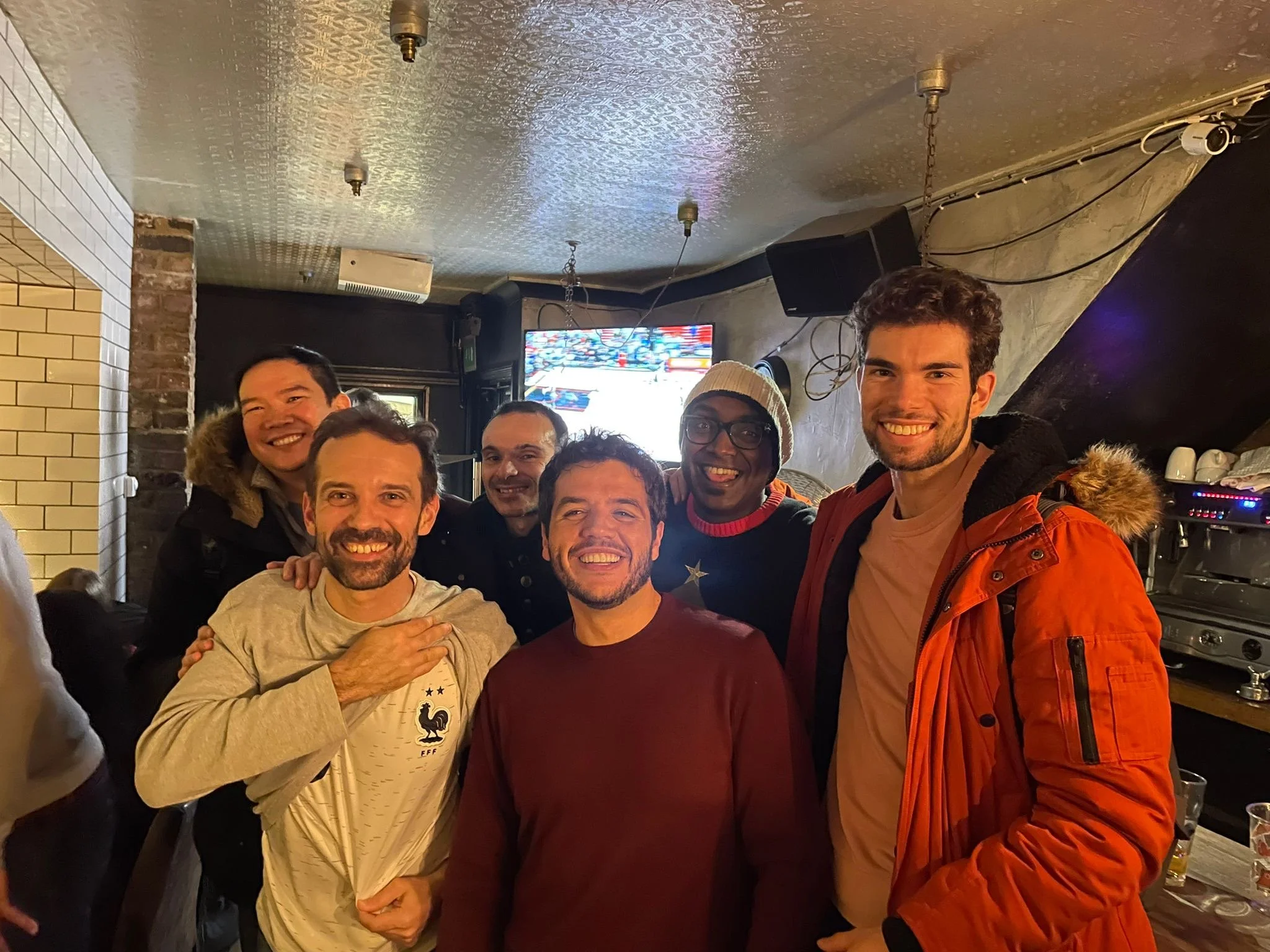 Group of six men smiling and posing for a photo inside a bar or pub, with a TV screen, stereo equipment, and speakers in the background.