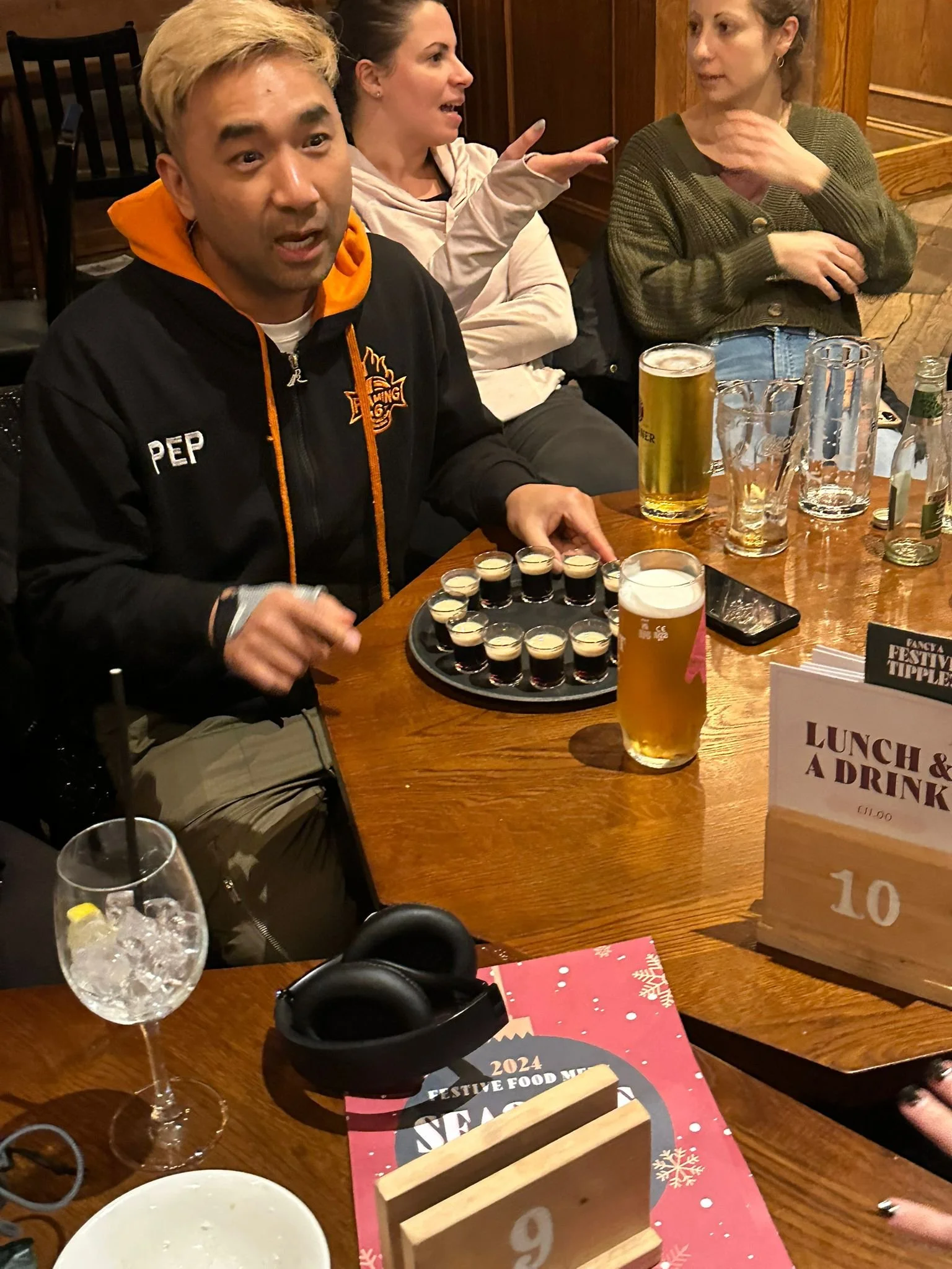 A group of people sitting at a wooden table in a restaurant or bar. One man in a black jacket with orange accents, labeled 'PEP,' is holding a tray of small shots. There are several glasses of beer on the table, along with a phone, a pair of black he