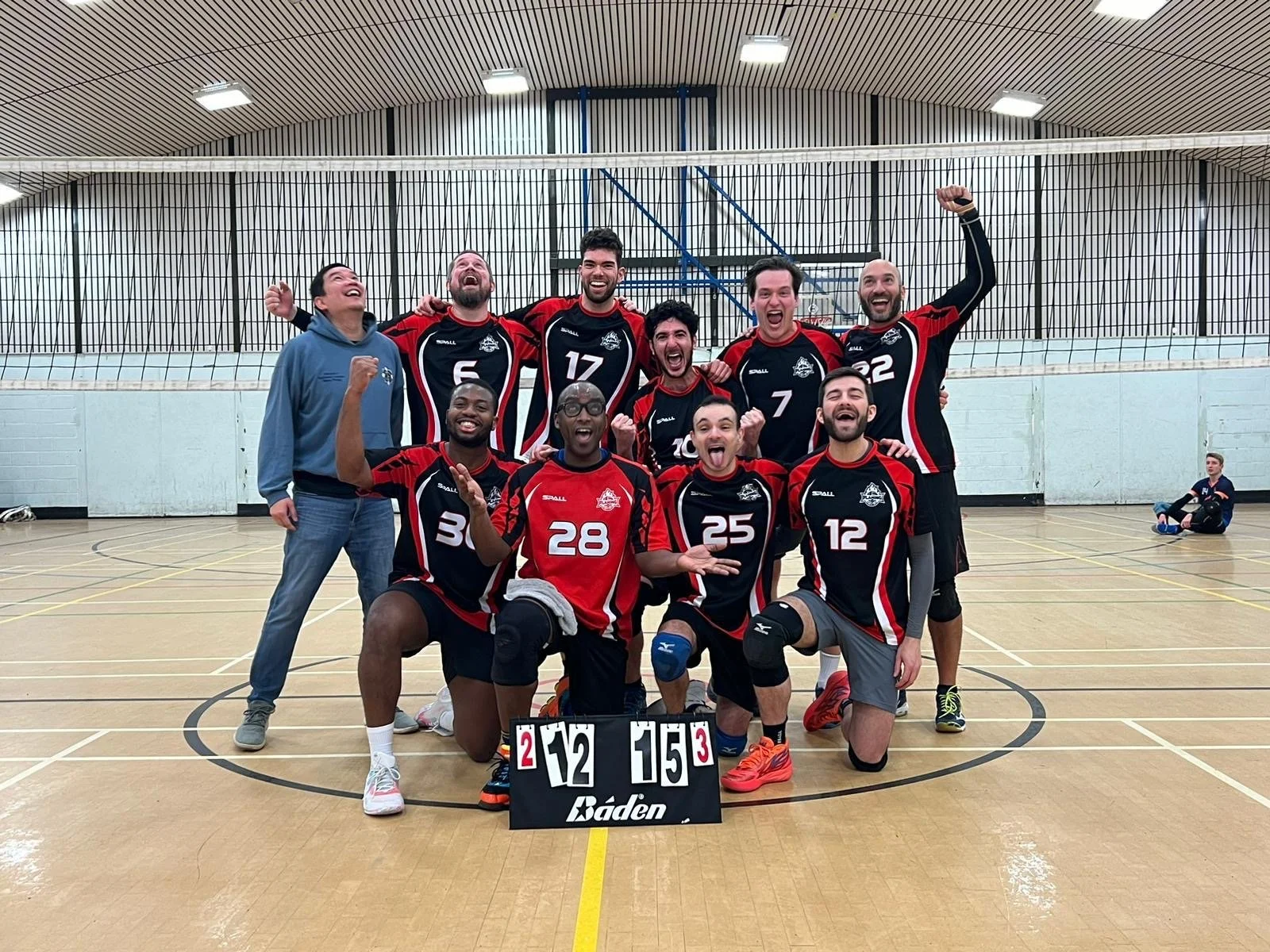 Indoor volleyball team group photo with players celebrating, some kneeling in front and others standing, in a gymnasium with volleyball net and scoreboard.