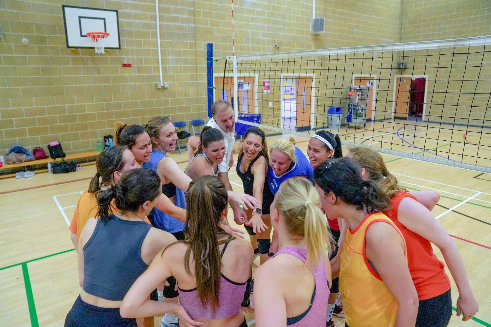 A group of women and a man huddle together smiling and cheering inside a gym on a volleyball court.