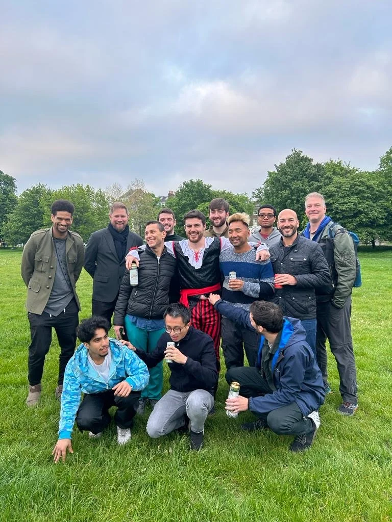 A group of 13 men gathered outdoors on a grassy field, some holding drinks, posing for a photo under a cloudy sky with trees and buildings in the background.