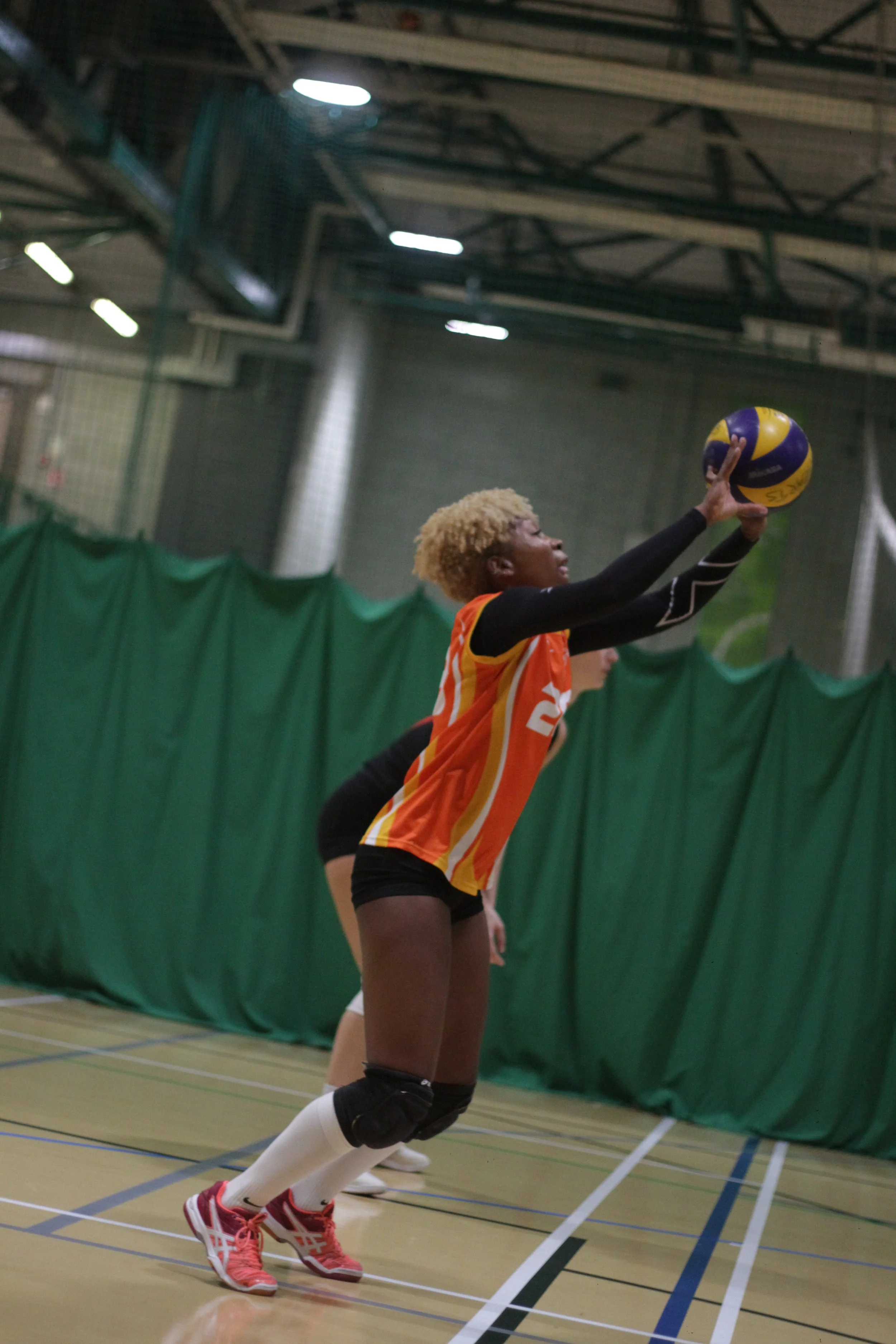 A female volleyball player with blonde curly hair wearing an orange and yellow jersey, black shorts, knee pads, and red shoes, is diving to set a blue and yellow volleyball during a match indoors.