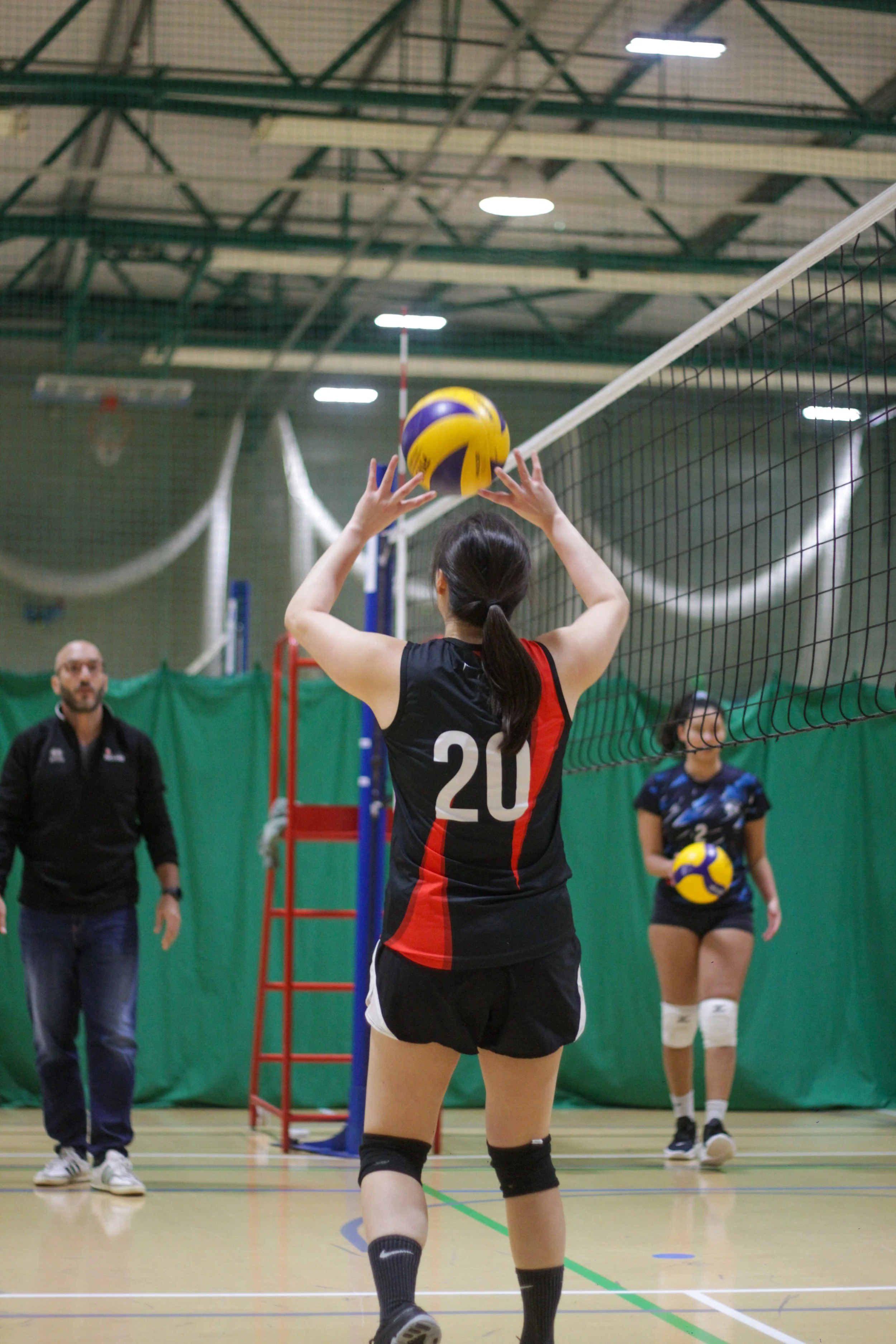 A female volleyball player with jersey number 20 is preparing to set a volleyball during a game in an indoor gymnasium, with a coach and another player in the background.