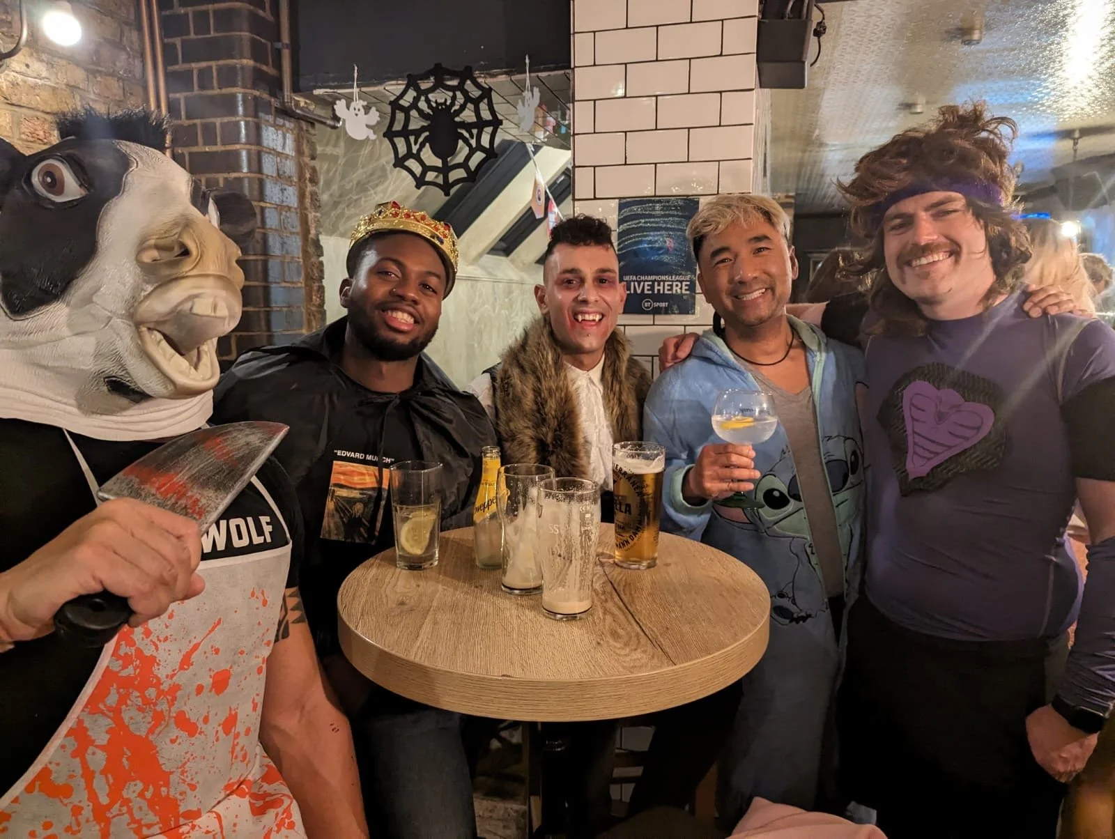 A group of five friends gathered around a high table at a pub celebrating, with one person wearing a wolf mask and holding a knife, all smiling and raising glasses of beer.