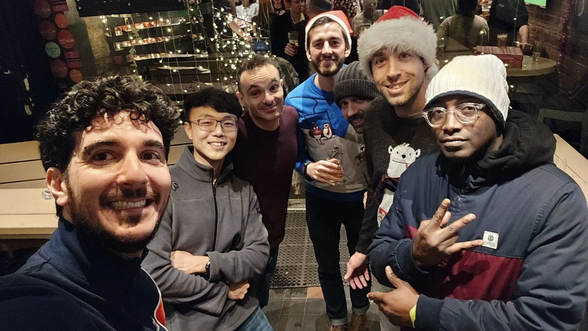 Group of seven friends celebrating Christmas at a festive indoor gathering, some wearing Santa hats and holiday sweaters, with Christmas decorations and lights in the background.