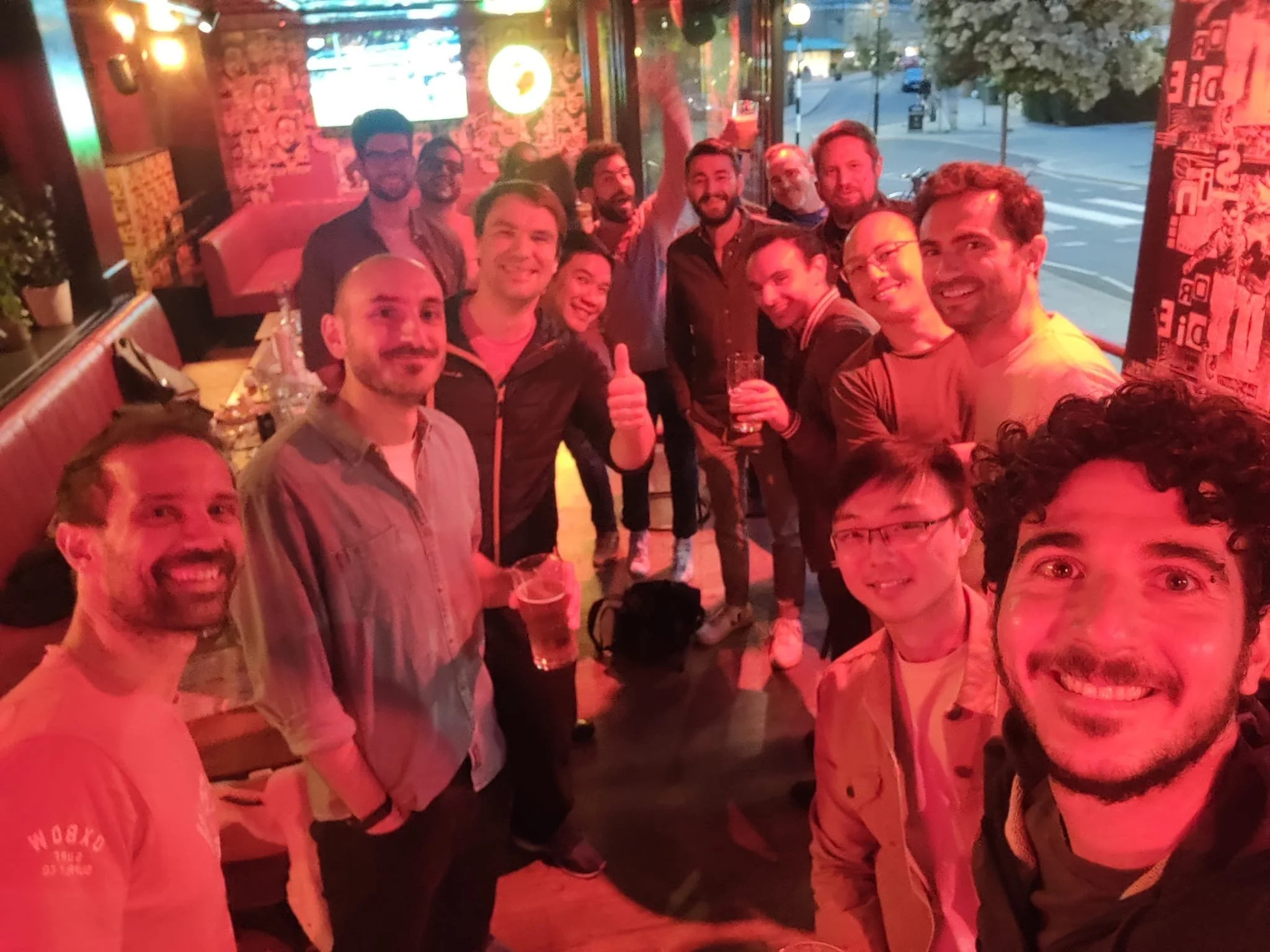 A group of people at a social gathering in a bar, smiling and posing for a selfie, with some holding drinks. The bar has a colorful, lively atmosphere with warm lighting and decor, and some outdoor scenery visible through windows.