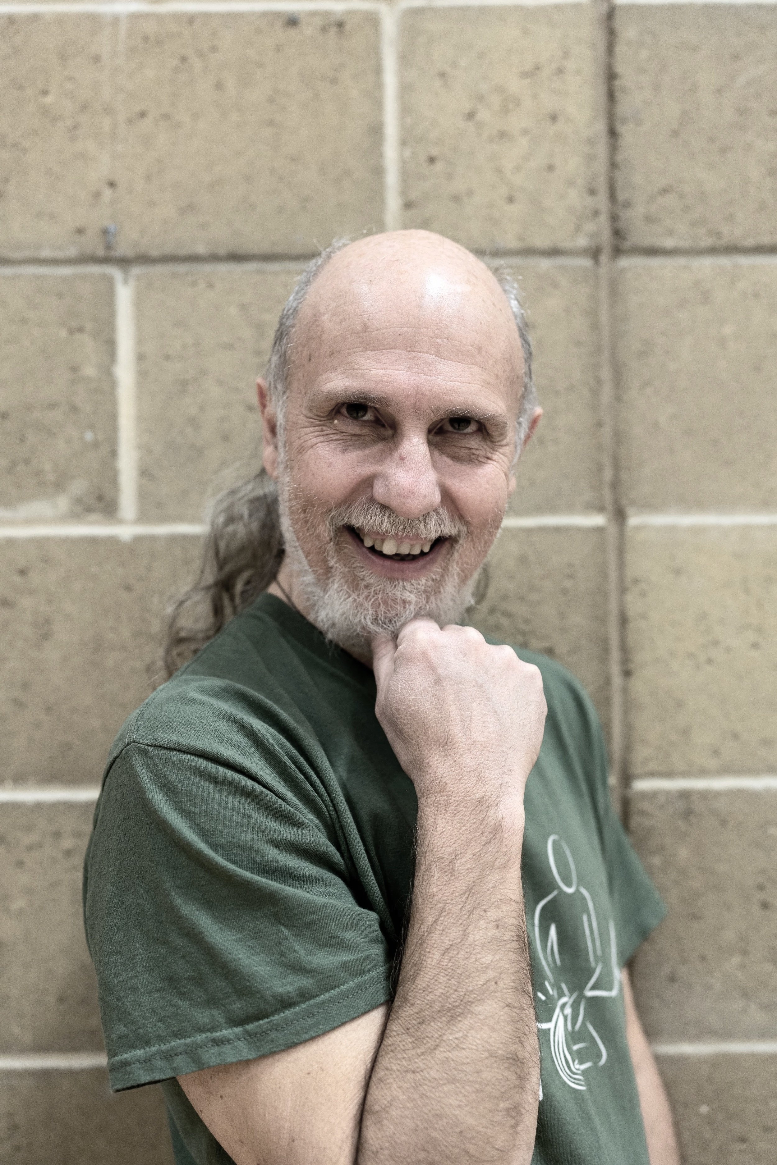 A smiling older man with a beard and no hair, wearing a green t-shirt, standing in front of a beige brick wall. He is touching his chin with his right hand, and appears cheerful.