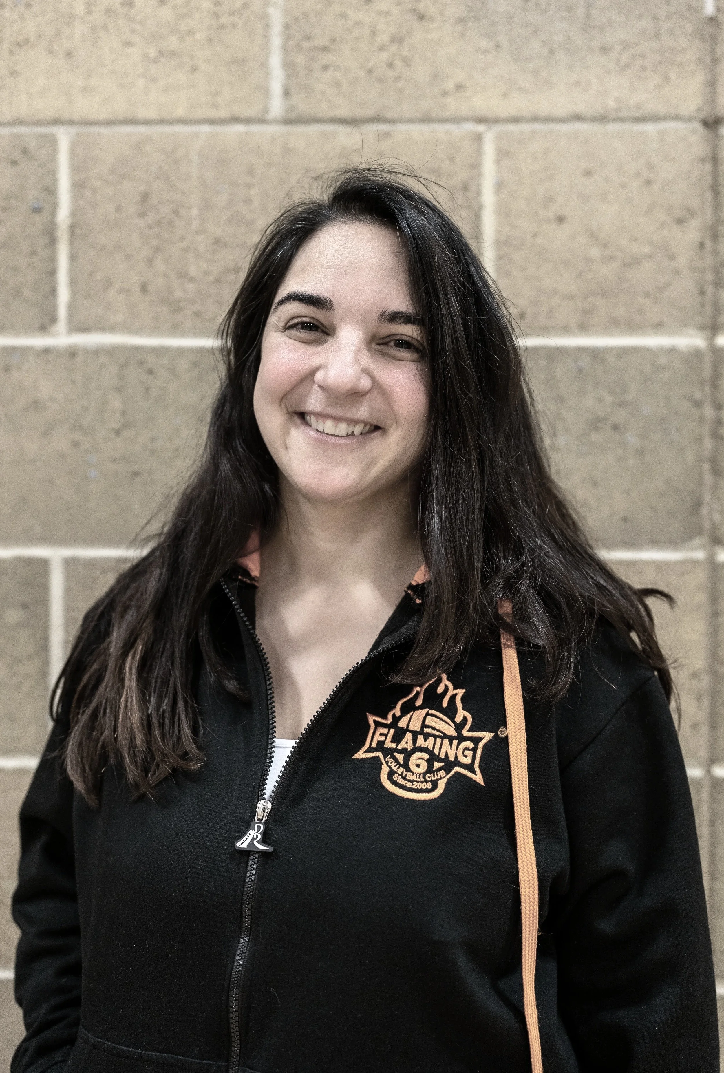 A woman with long dark hair smiling, wearing a black jacket with a flaming volleyball logo on the chest, standing in front of a beige brick wall.