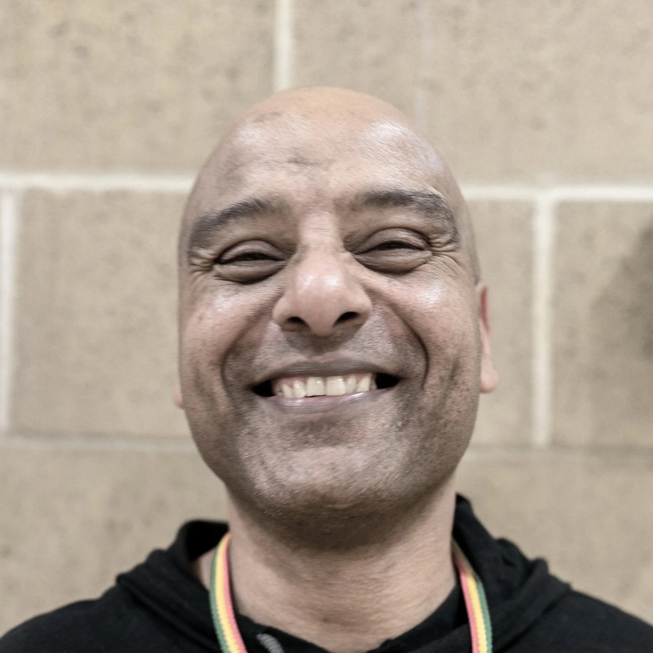Close-up of smiling man with shaved head, wearing a black hoodie and a rainbow-colored necklace in front of a beige brick wall.