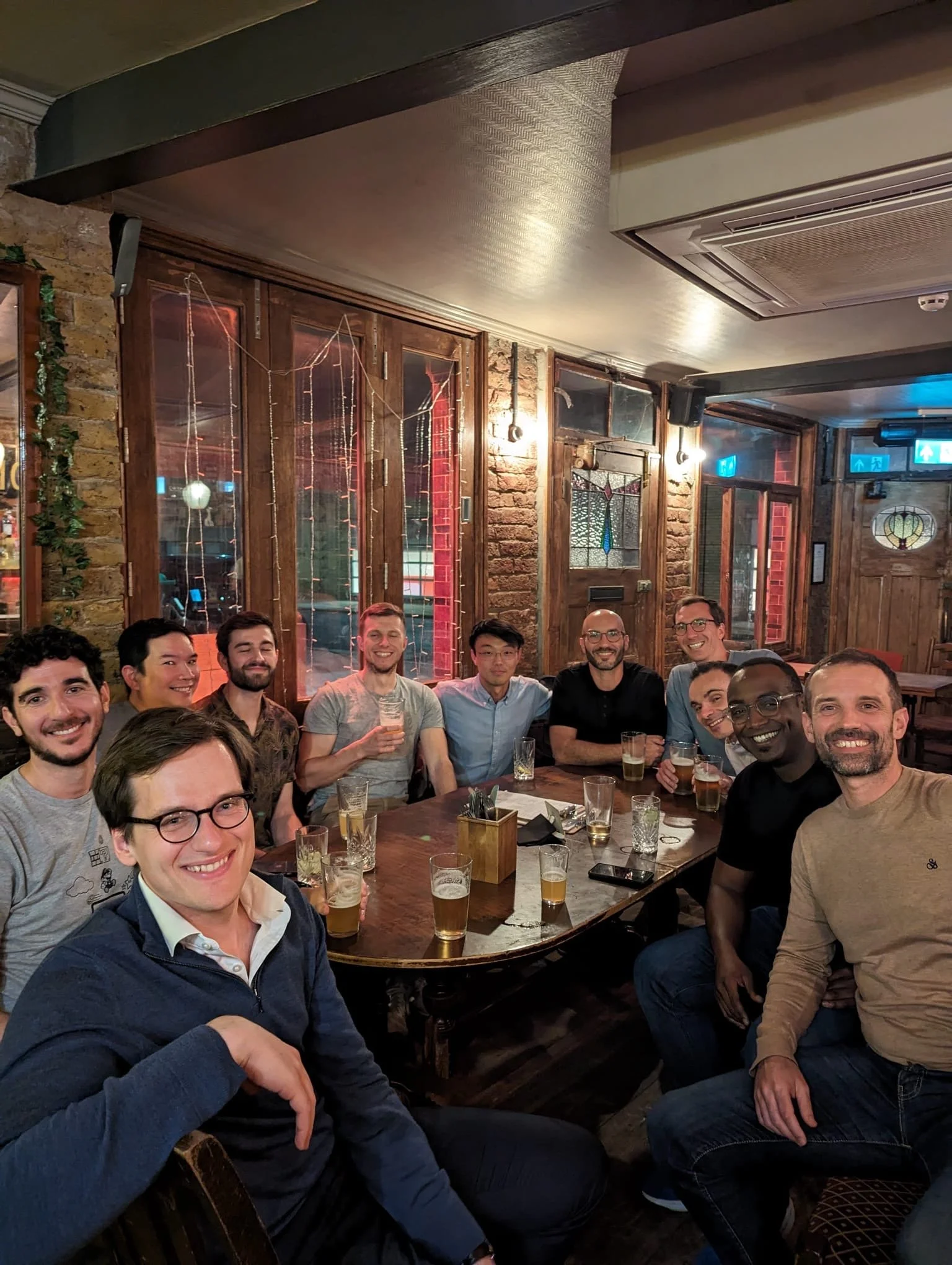 Group of people smiling and sitting around a table in a pub or restaurant, with drinks in front of them, warmly lit interior with wooden and brick decor.
