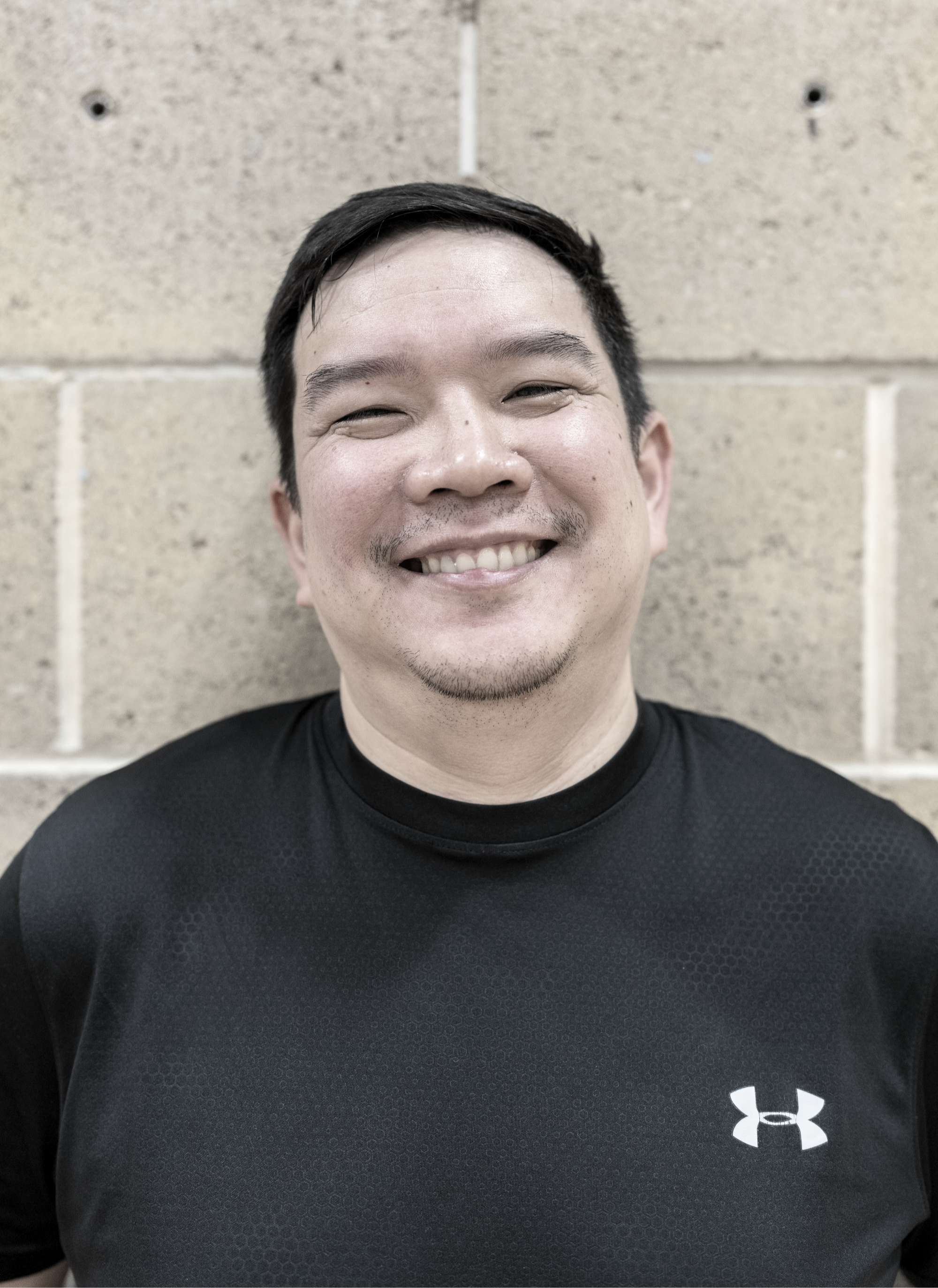 A smiling man with short dark hair, light facial hair, and a black Under Armour shirt standing in front of a beige brick wall.