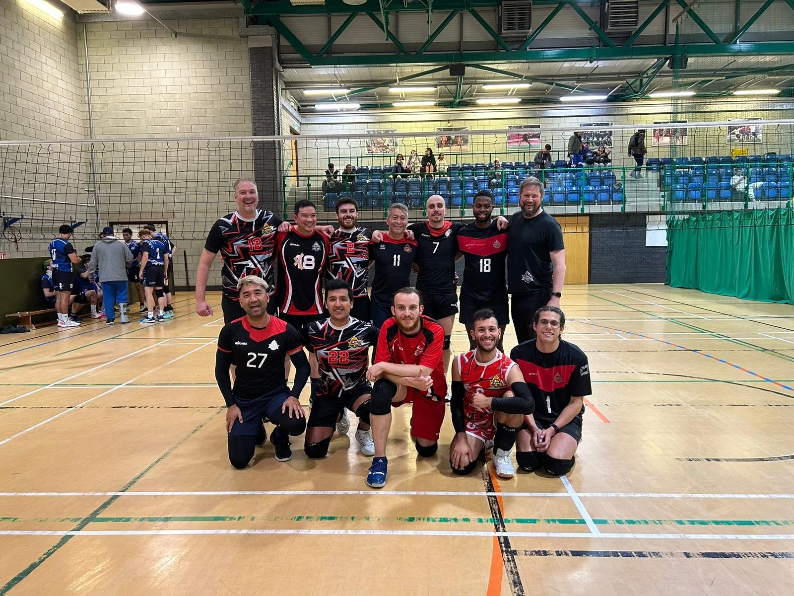 A group of eleven men in volleyball uniforms posing for a team photo on an indoor volleyball court. They are smiling, some kneeling in the front row and some standing in the back row. The court has a volleyball net, and there are spectators and other