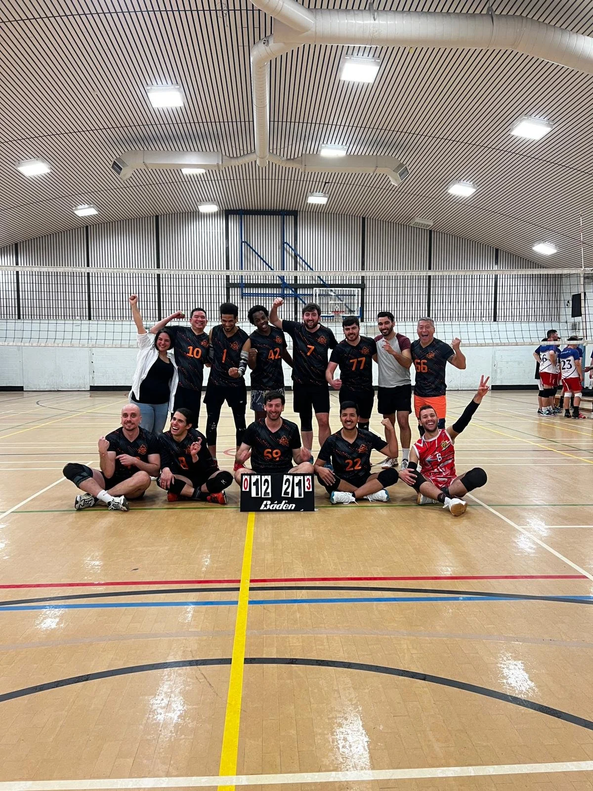 A volleyball team of 12 players posing for a photo in an indoor gymnasium. The team is holding a scoreboard showing a score of 12 to 25, with 3 sets played. The players are celebrating, some with fists in the air and others making peace signs. The gy
