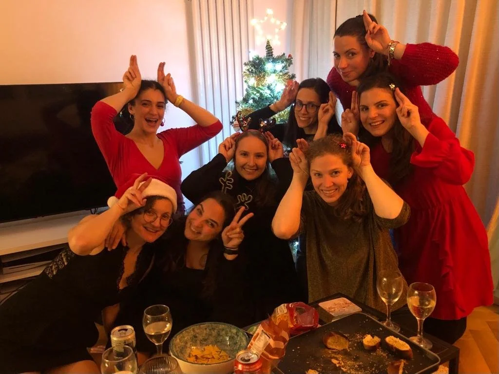 A group of eight women celebrating around a table with snacks and drinks, wearing festive Christmas attire and posing with playful gestures in front of a decorated Christmas tree.