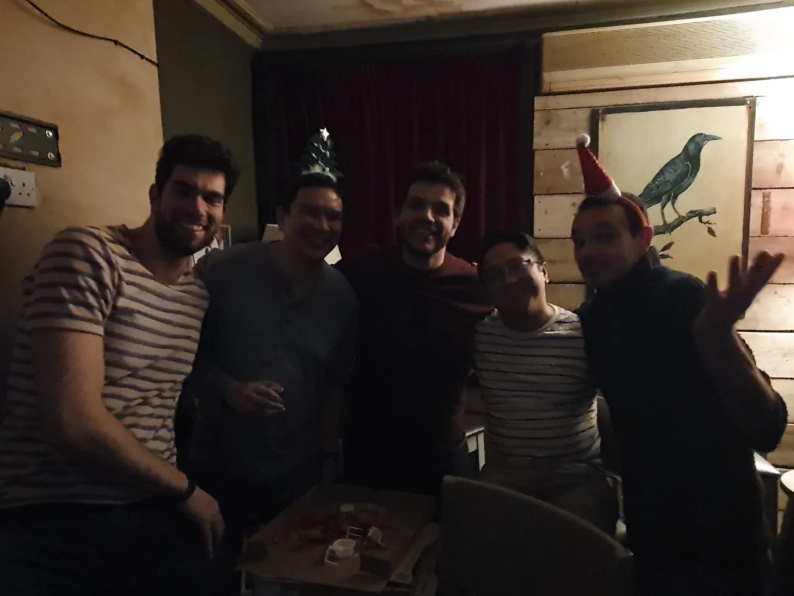 Group of five men smiling at a party in a dimly lit room, one wearing a Santa hat with a bird decoration, celebrating.