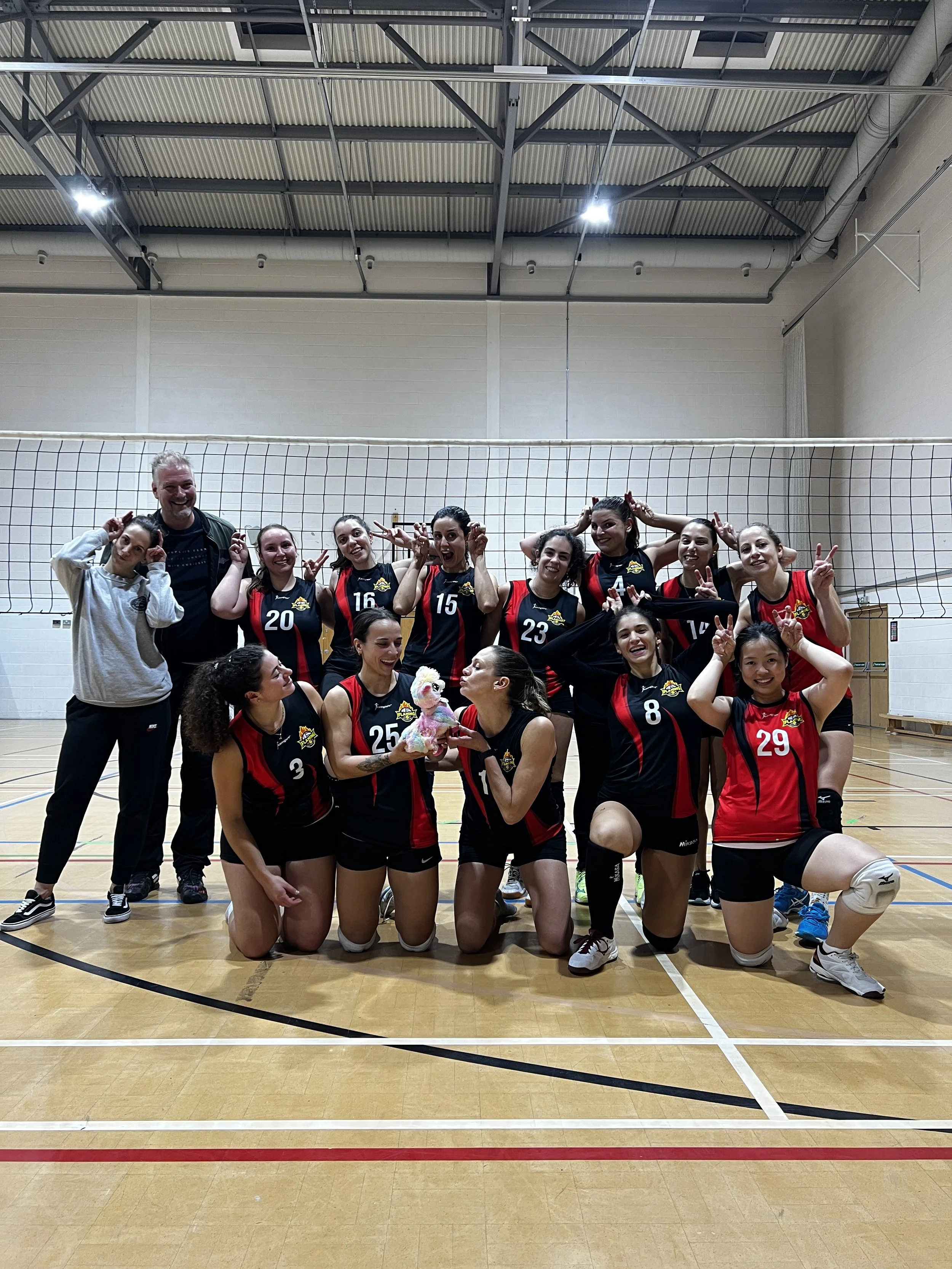 A women's volleyball team celebrating on an indoor court with their coach, holding a plush toy and making peace signs.