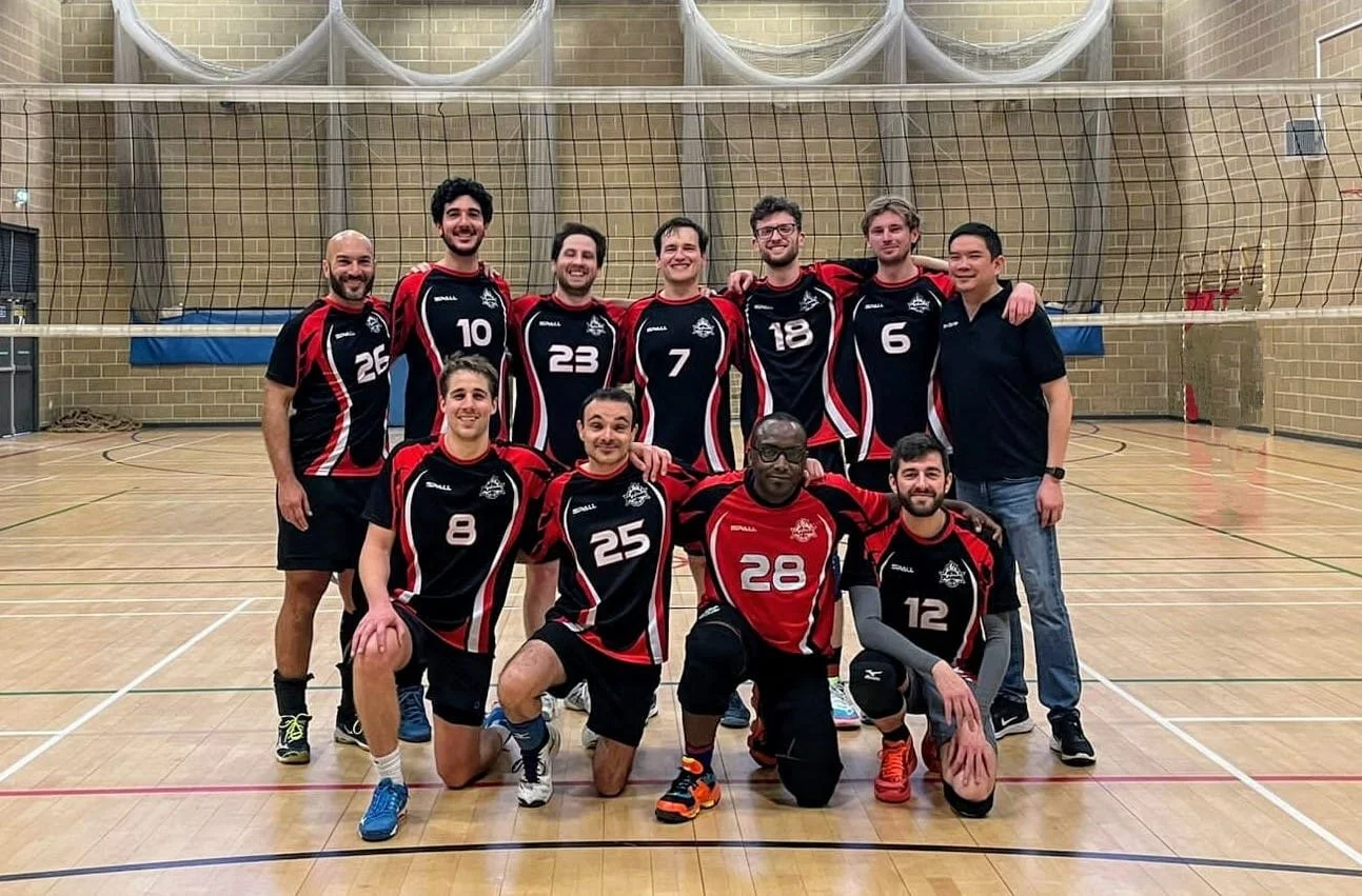 A group of eleven male volleyball players in black and red uniforms celebrating in an indoor gymnasium, with a volleyball net behind them.
