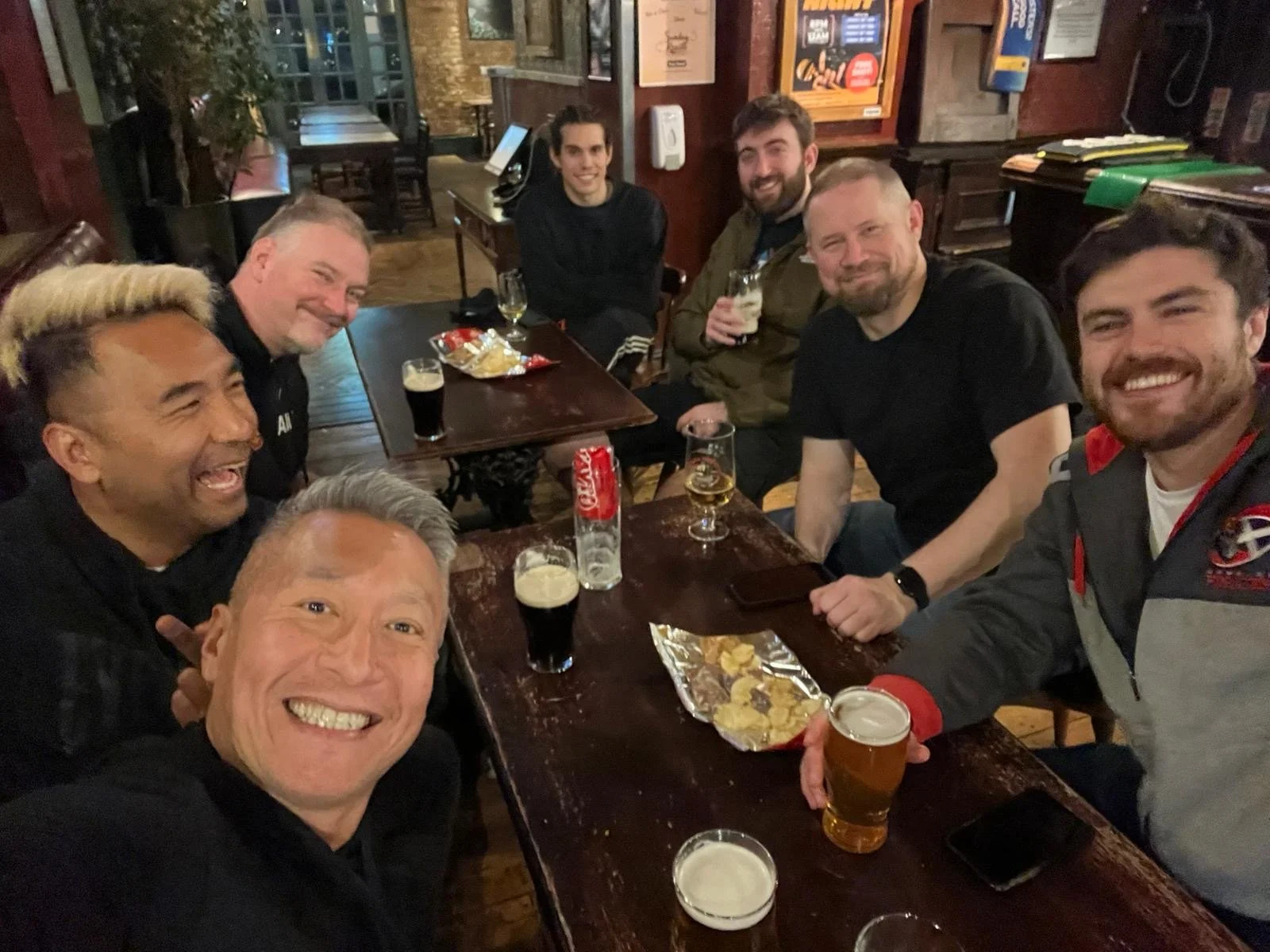 Group of eight friends smiling and posing for a photo at a wooden table in a dimly lit pub, with drinks and snacks, celebrating together.