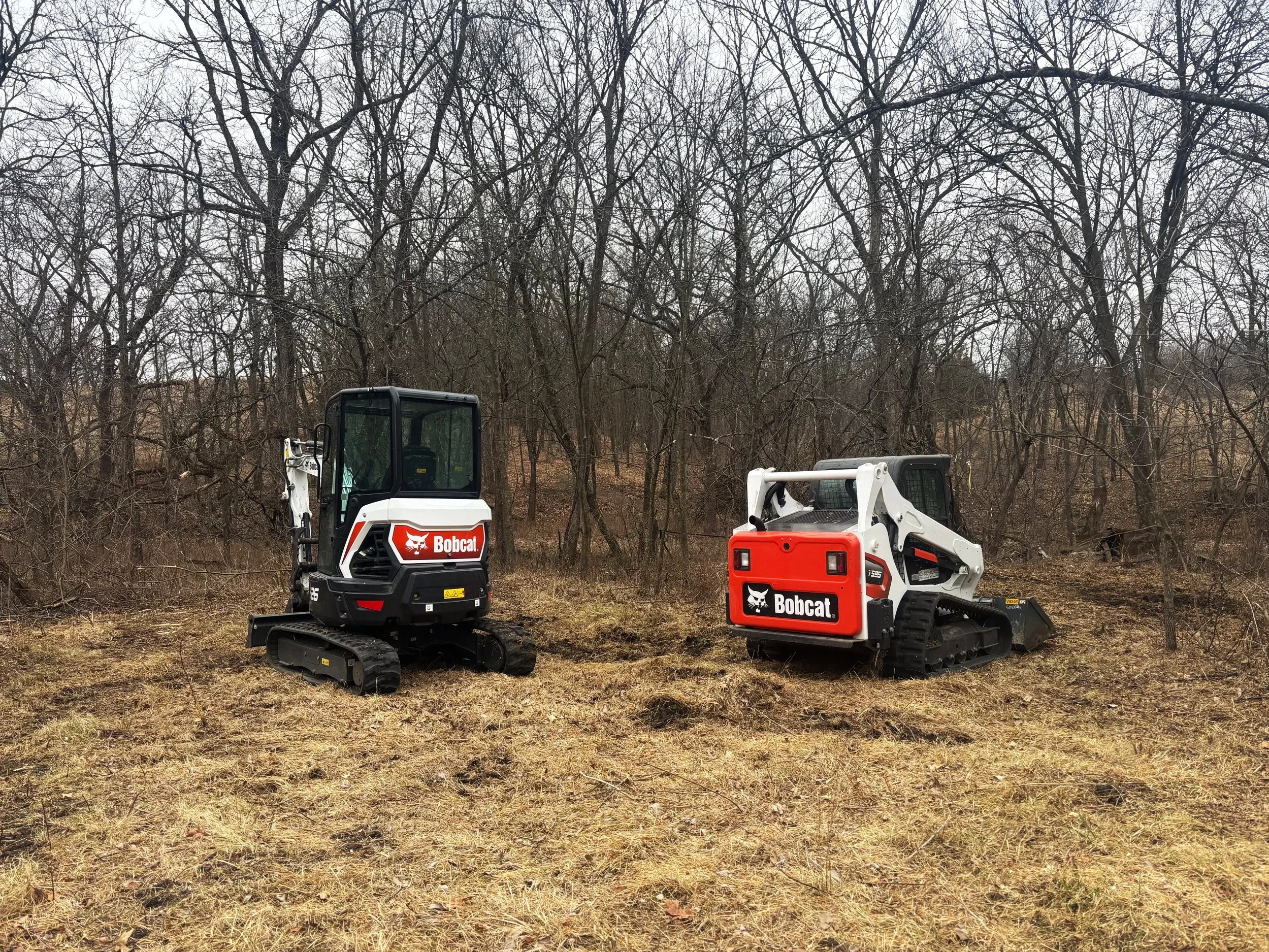 Two compact Bobcat construction vehicles, one black and white, parked outdoors on grass with leafless trees in the background.