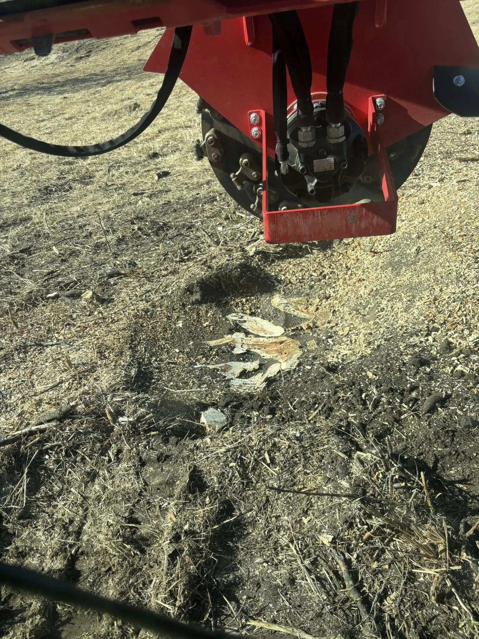 Close-up of a red agricultural machine attachment with hydraulic hoses, positioned over a patch of dry, charred soil with remnants of burnt plant material