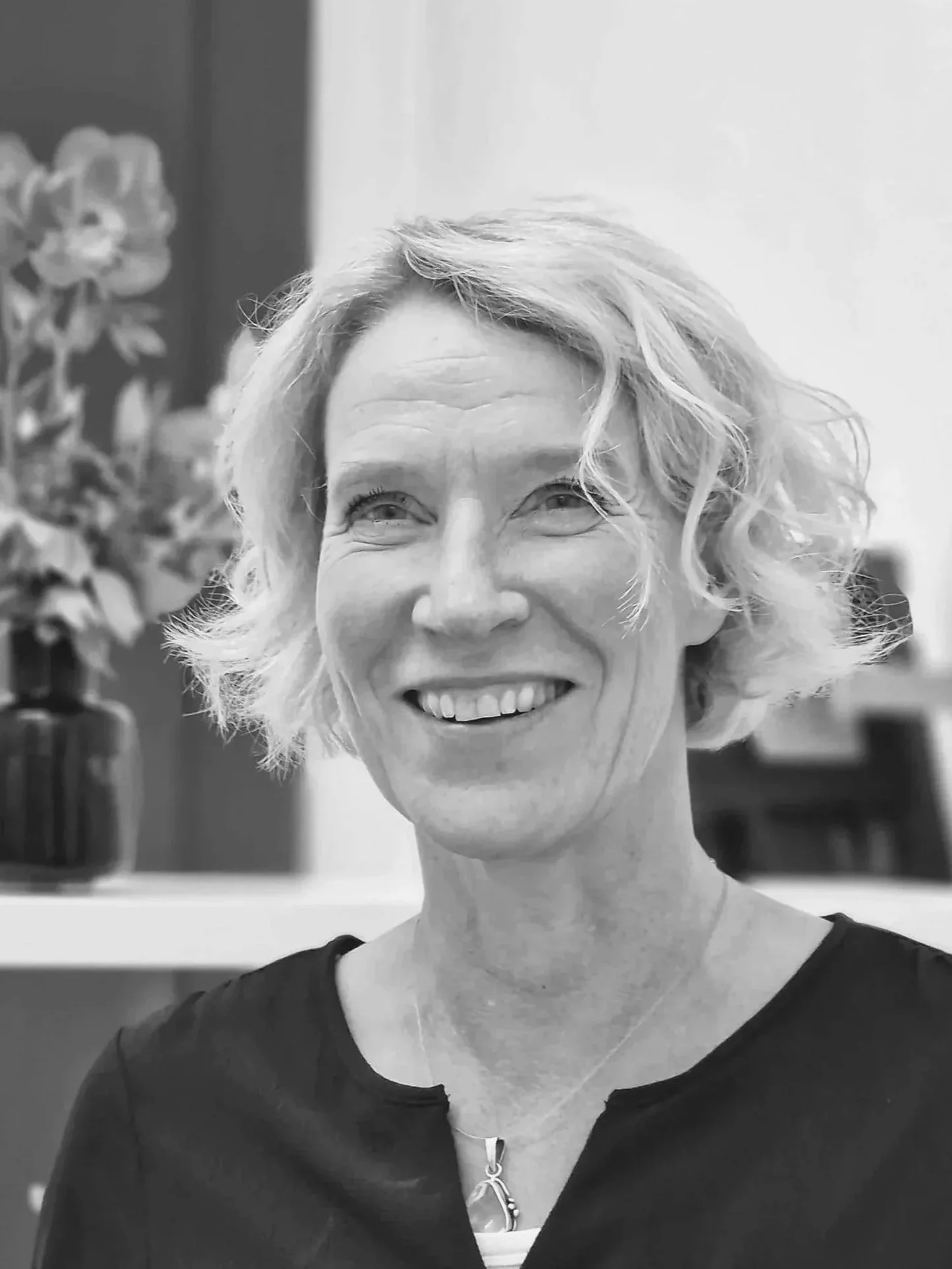 Schwarz-weiß Portrait einer lächelnden Frau mit kurzen, lockigen Haaren, trägt ein dunkles Oberteil und eine Halskette, im Hintergrund sind Blumen und ein Bücherregal zu sehen.