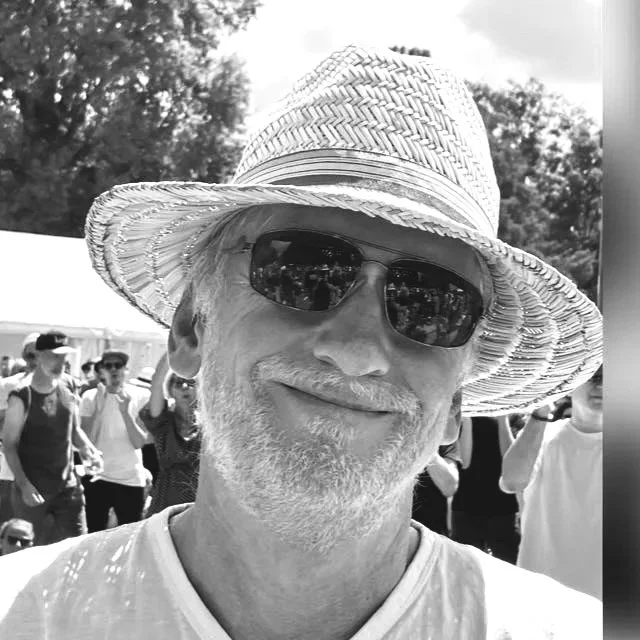 Ein älterer Mann mit weißem Bart, Sonnenbrille und Strohhut, lächelnd auf einem Festival oder einem öffentlichen Anlass, im Hintergrund Menschen und Bäume.