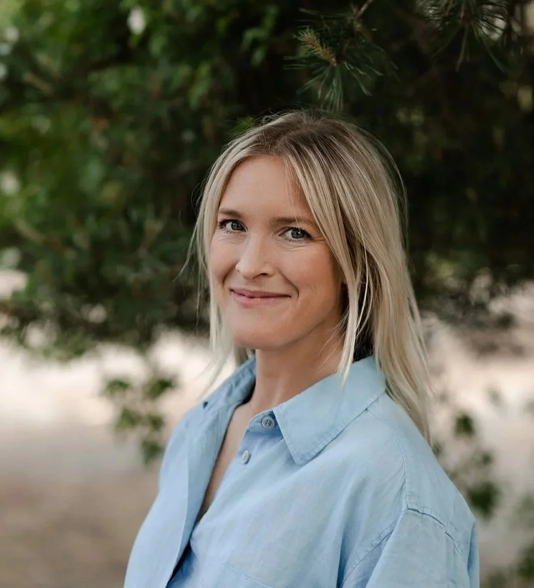 En leende kvinna med blont hår som bär en ljusblå skjorta, står framför tallar i utomhusmiljö.